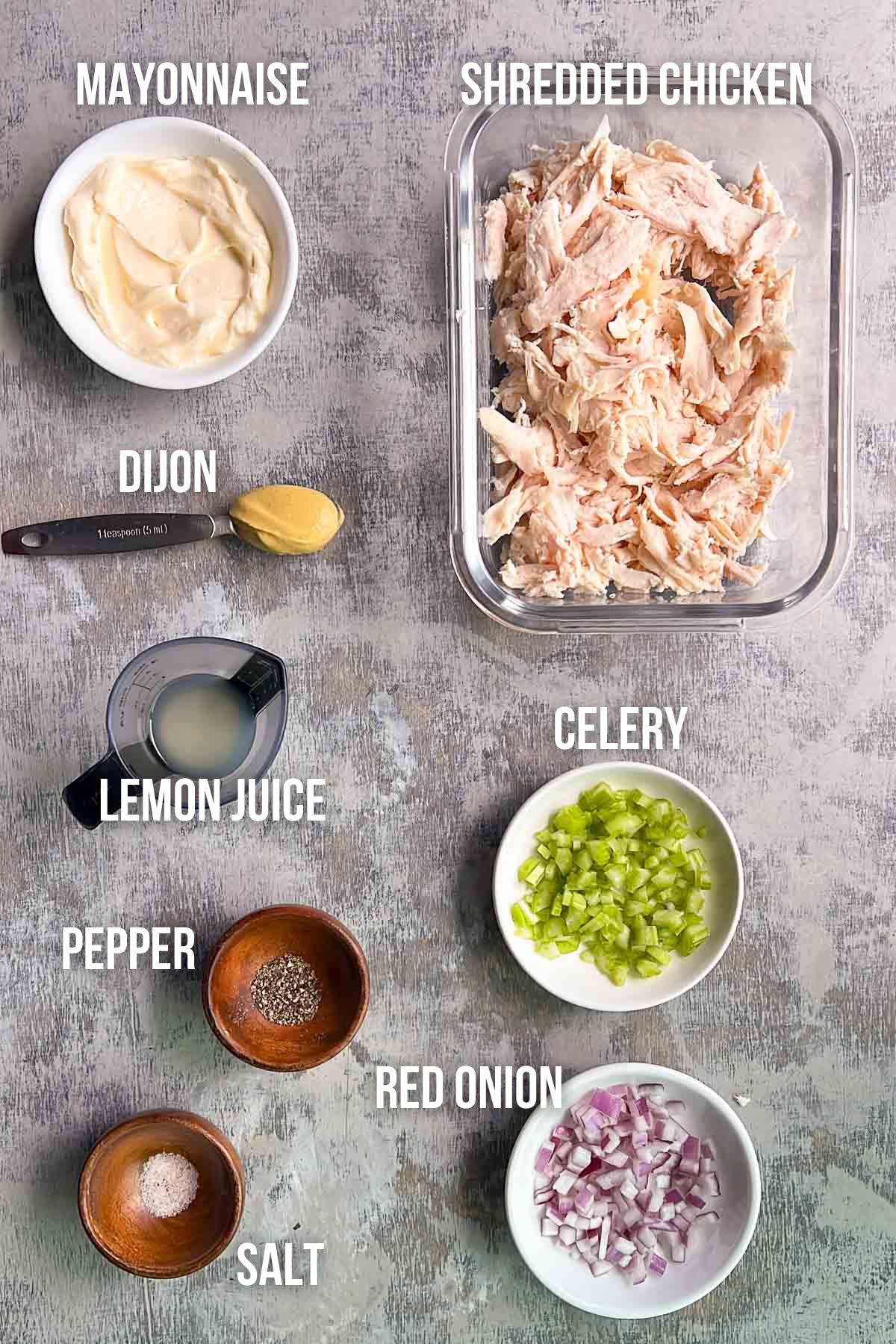 Ingredients for chicken salad displayed on a gray surface: mayonnaise, shredded chicken, Dijon mustard, lemon juice, celery, red onion, pepper, and salt in separate bowls.