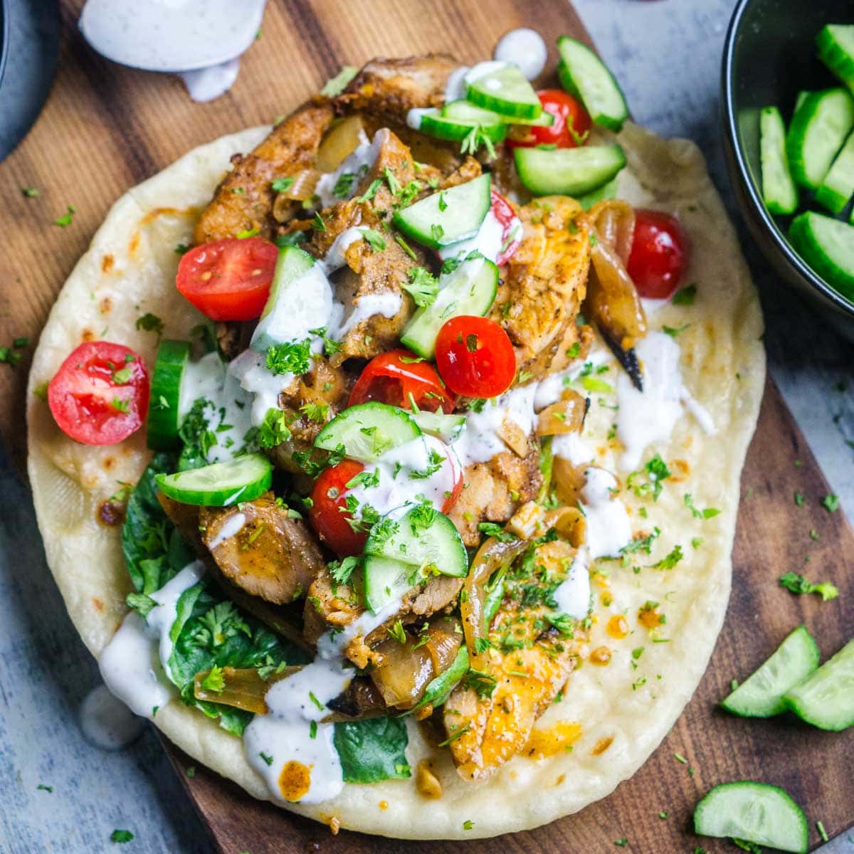 Flatbread topped with chicken shawarma, sliced cucumbers, cherry tomatoes, greens, onions, fresh herbs, and drizzled with a white sauce, served on a wooden board.