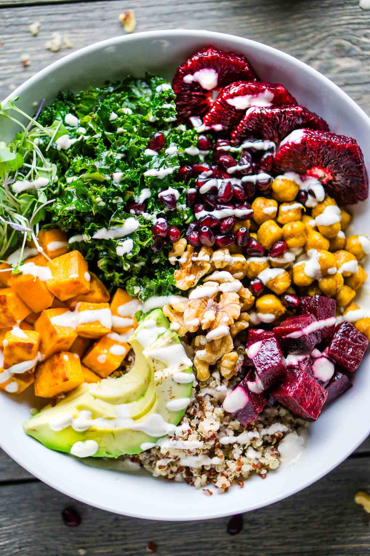 Buddha bowl ingredients consisting of kale, quinoa, diced sweet potatoes, chickpeas, blood oranges, diced beets, walnuts, microgreens, pomegranate arils, and avocado with drizzled Buddha bowl sauce in white bowl