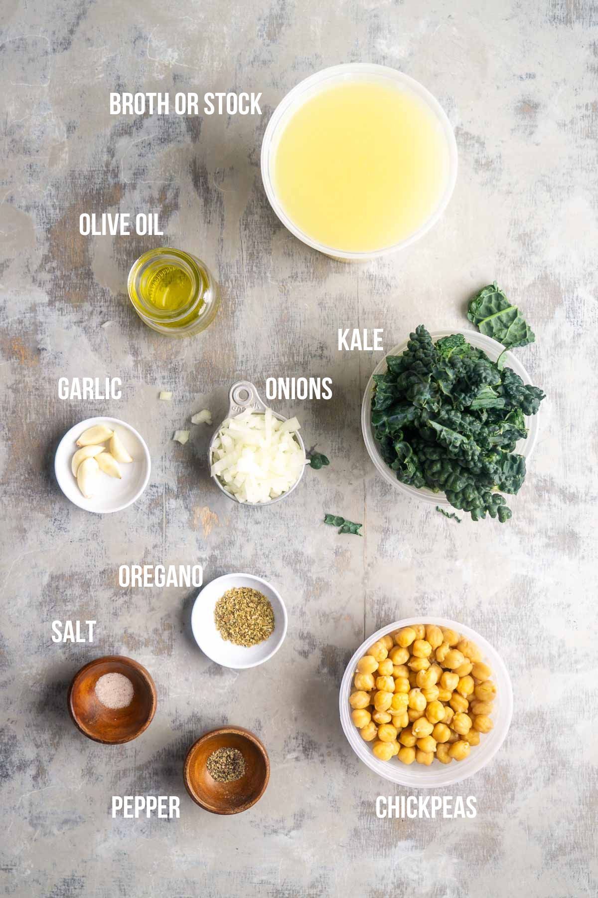 Top-down view of labeled chickpea kale soup ingredients on a gray surface: broth or stock, olive oil, garlic, oregano, onions, kale, chickpeas, salt, and pepper.