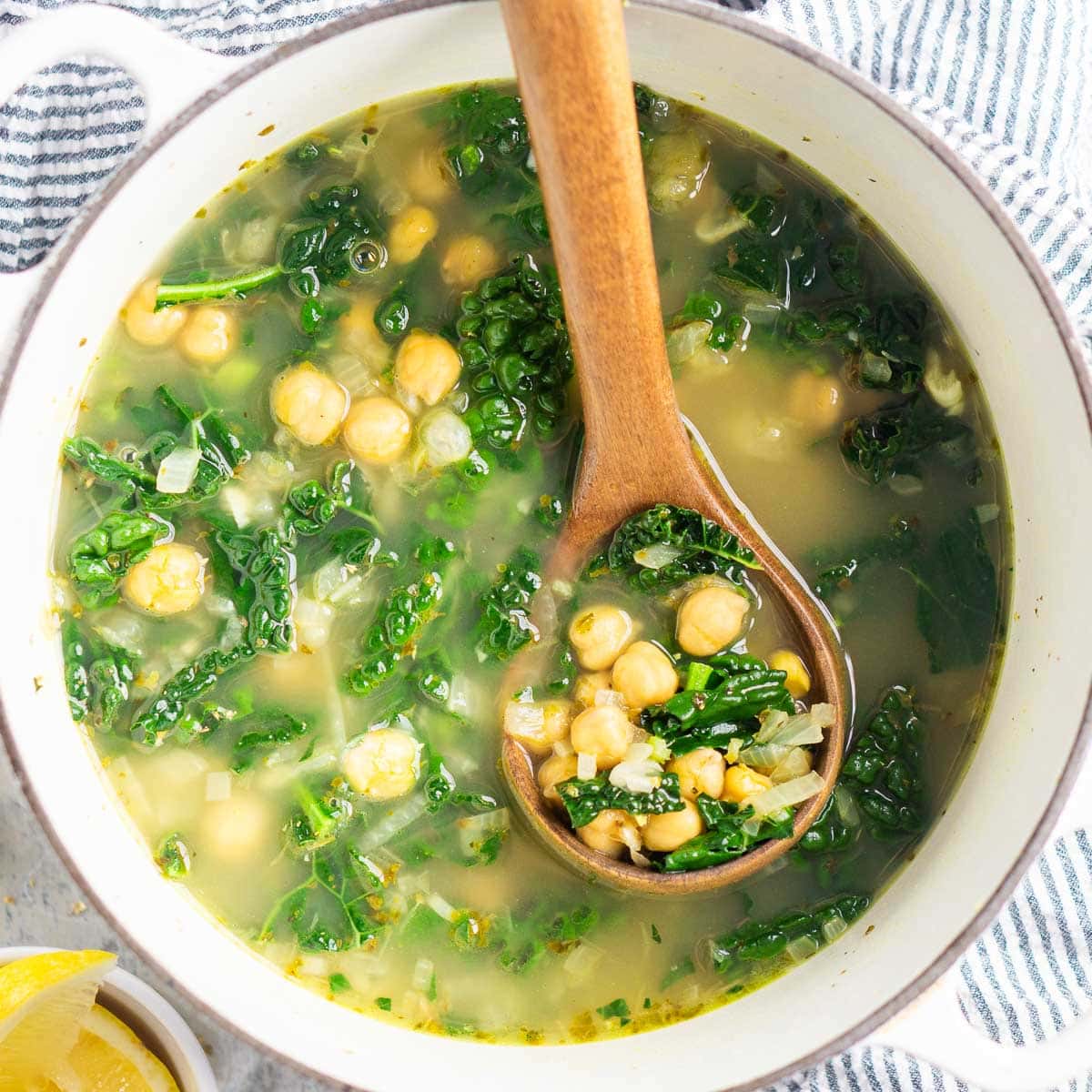 A pot of chickpea kale soup with a wooden spoon, featuring clear broth and visible chickpeas and kale. A bowl of lemons is nearby.