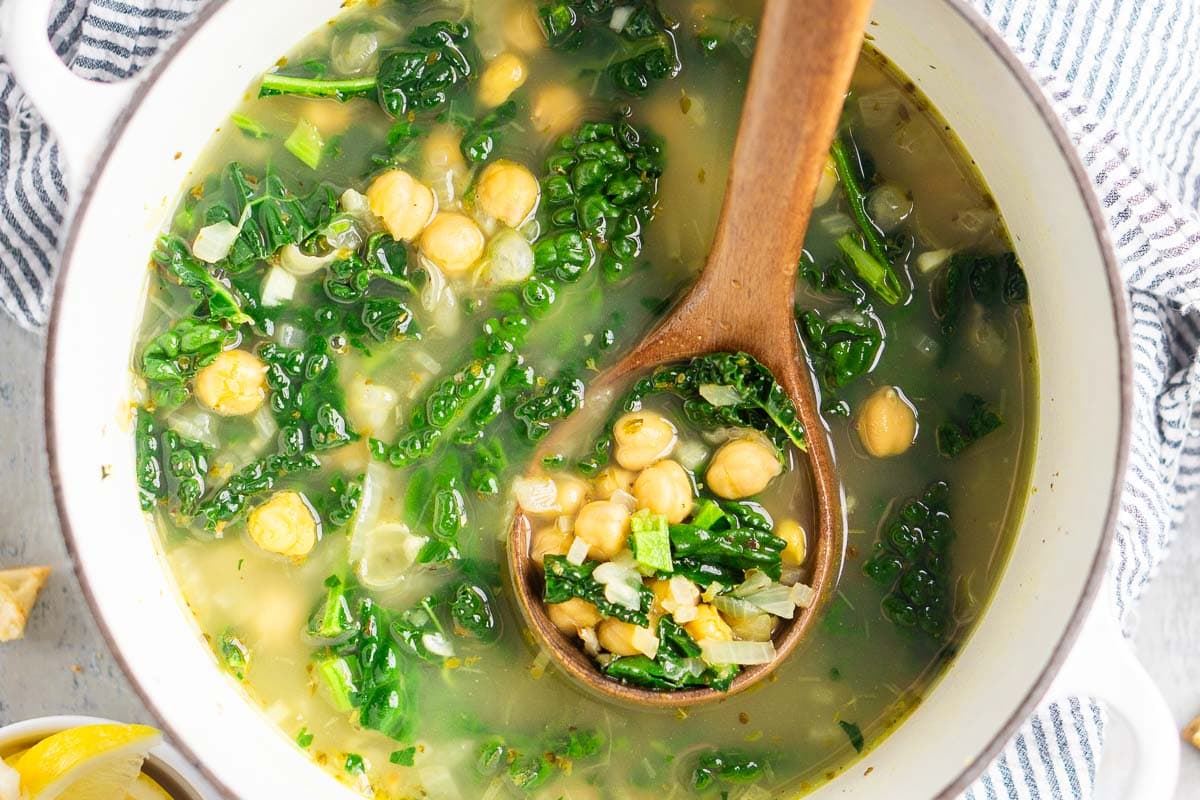 A pot of chickpea kale soup with a wooden spoon, featuring clear broth and visible chickpeas and kale. A bowl of lemons is nearby.