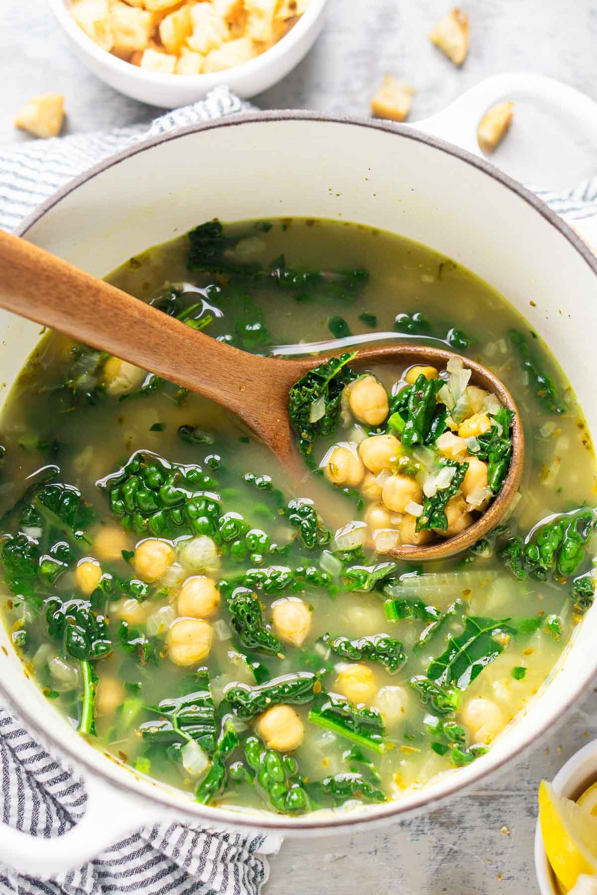 A pot of chickpea kale soup with a wooden spoon, featuring clear broth and visible chickpeas and kale. A bowl of croutons is nearby.