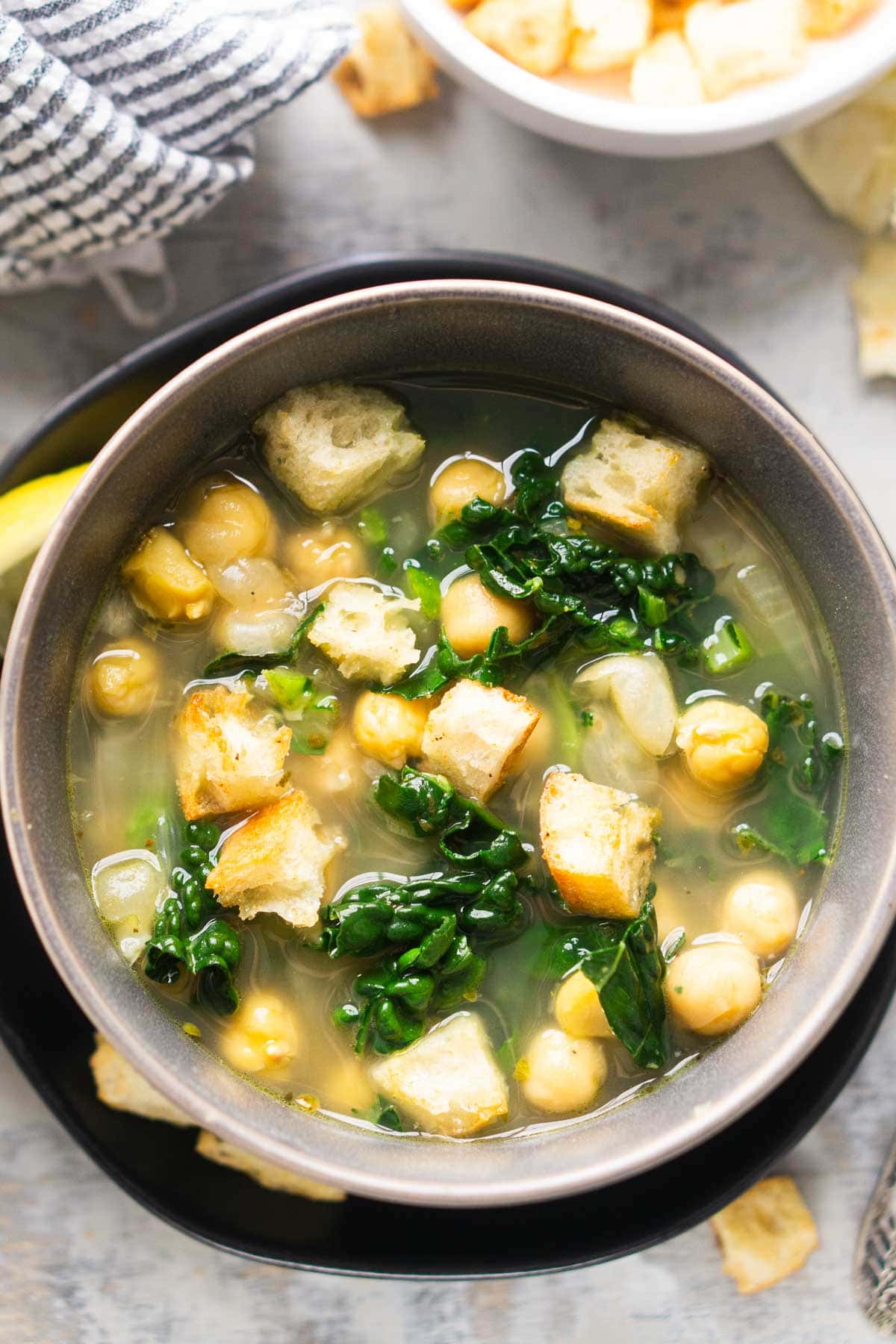 A bowl of soup with chickpeas, kale, and croutons, garnished with a lemon wedge, sits on a black plate next to a bowl of extra croutons.