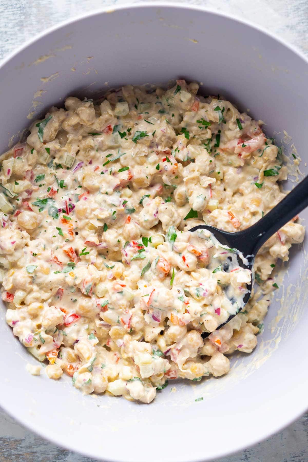 A mixing bowl filled with creamy chickpea salad, featuring chopped vegetables and fresh herbs, with a black spoon resting inside.