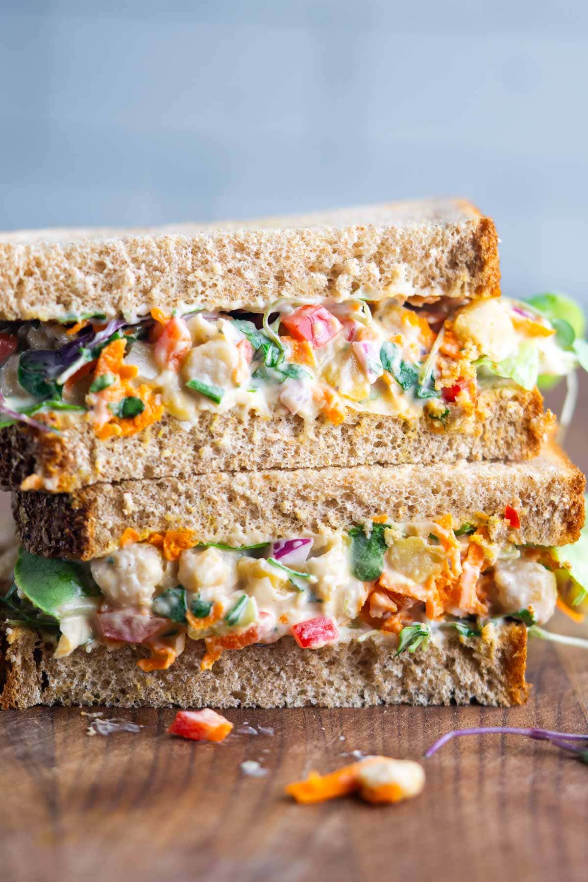 Two stacked sandwich halves on whole grain bread, filled with a colorful chickpea salad on a cutting board.