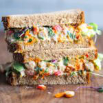 Two stacked sandwich halves on whole grain bread, filled with a colorful chickpea salad on a cutting board.