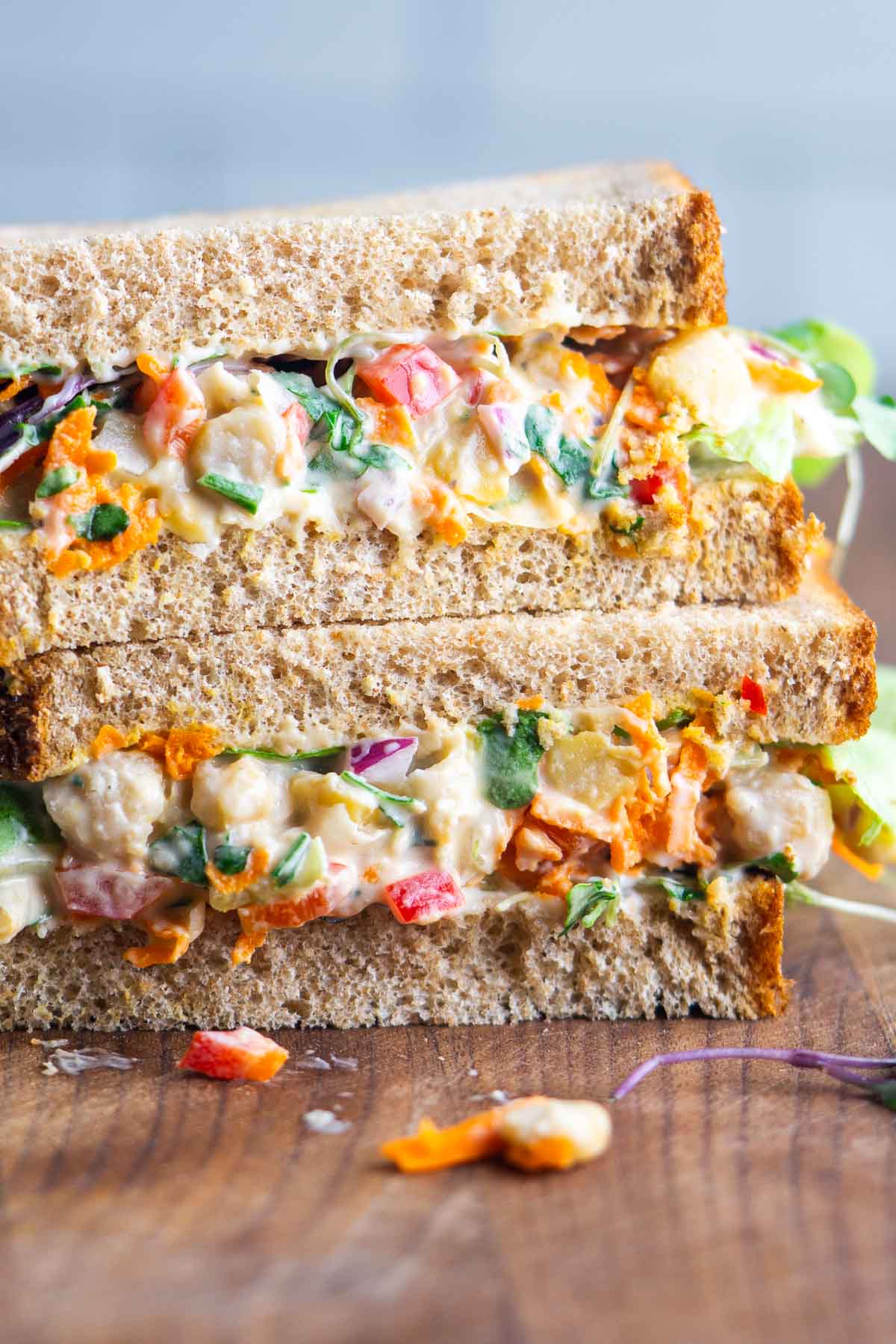 Two stacked sandwich halves on whole grain bread, filled with a colorful chickpea salad on a cutting board.