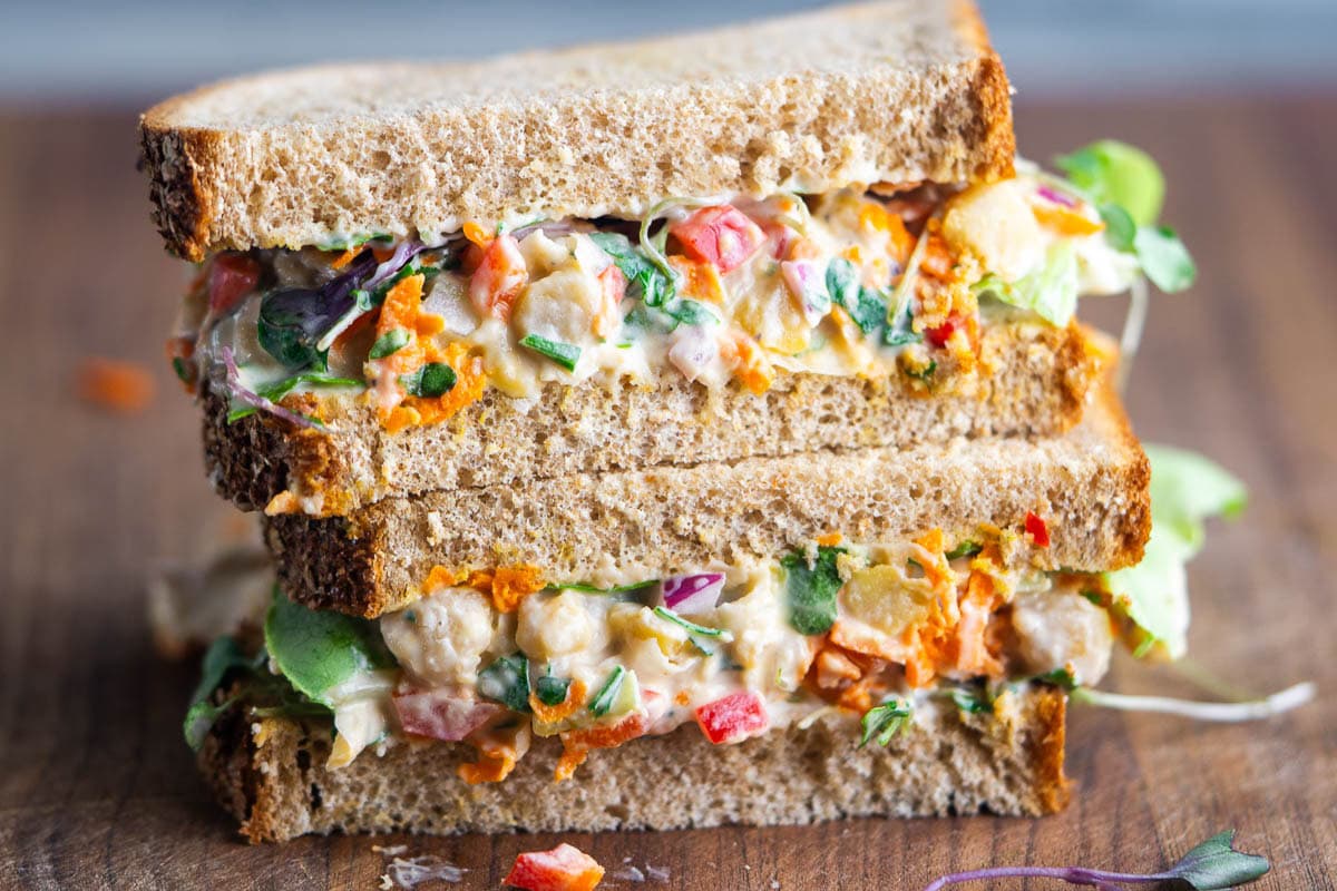 Two stacked sandwich halves on whole grain bread, filled with a colorful chickpea salad on a cutting board.