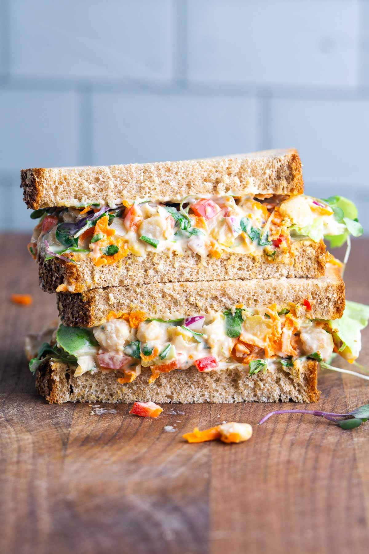 Two stacked sandwich halves on whole grain bread, filled with a colorful chickpea salad on a cutting board.