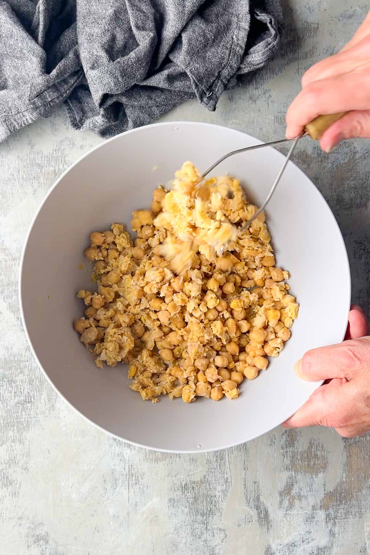 Hands using a potato masher to mash chickpeas in a gray bowl on a light-colored surface with a gray cloth nearby.