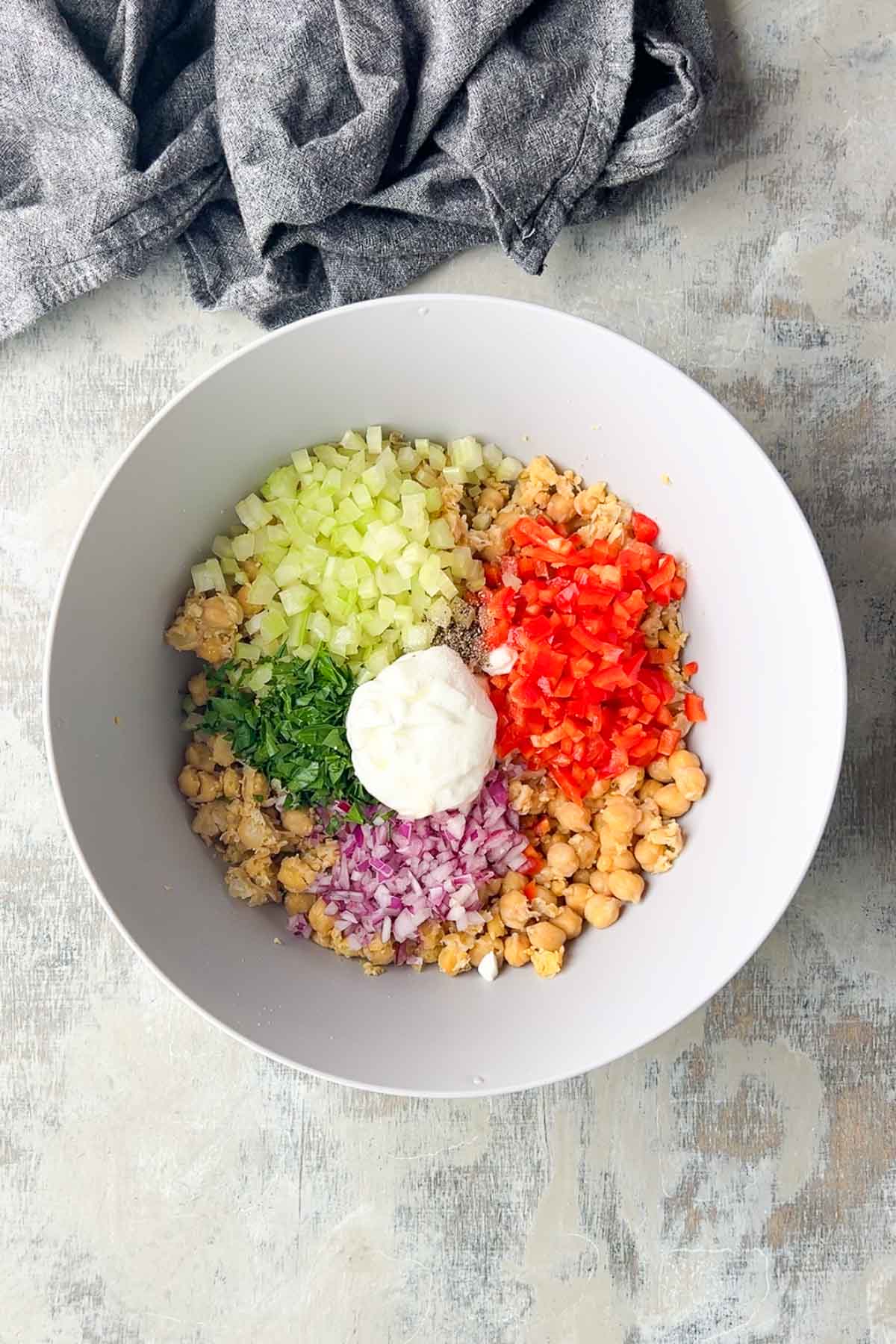 A white bowl contains chopped chickpeas, celery, red pepper, red onion, parsley, and a dollop of mayonnaise on a light-colored surface with a gray cloth nearby.