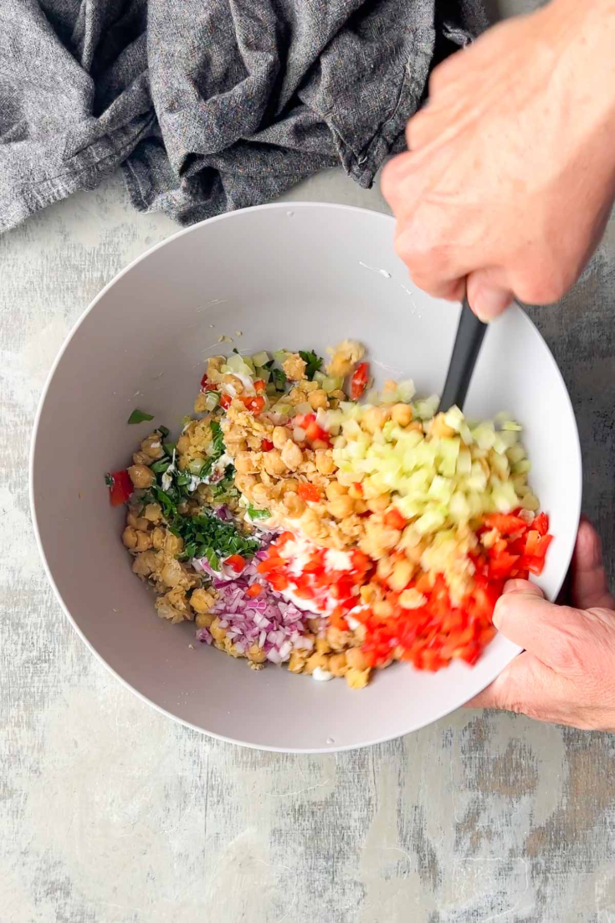 A person mixes chopped vegetables and chickpeas in a white bowl with a black spatula on a light-colored surface.