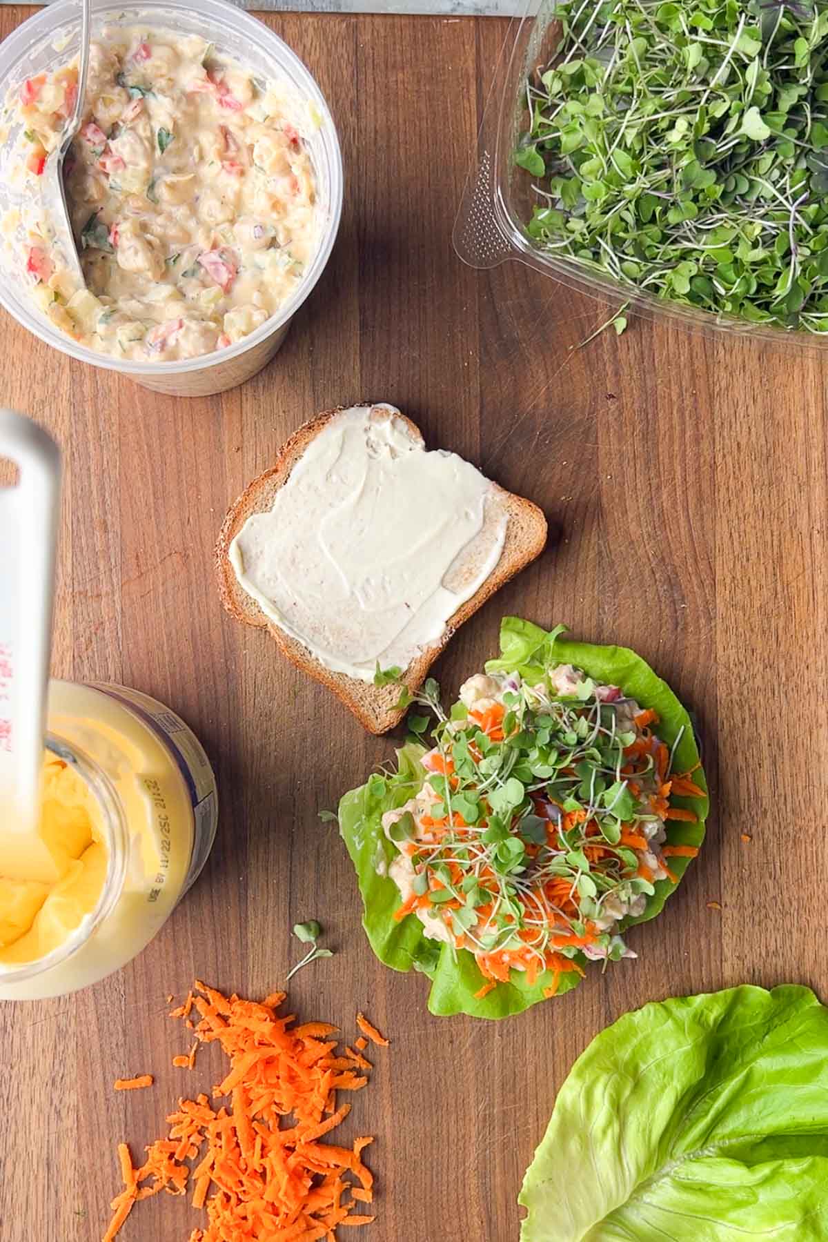Overhead view of sandwich prep with a slice of bread spread with mayonnaise, lettuce topped with chickpea salad, grated carrots, microgreens, and jars of salad and mayonnaise on a wooden surface.