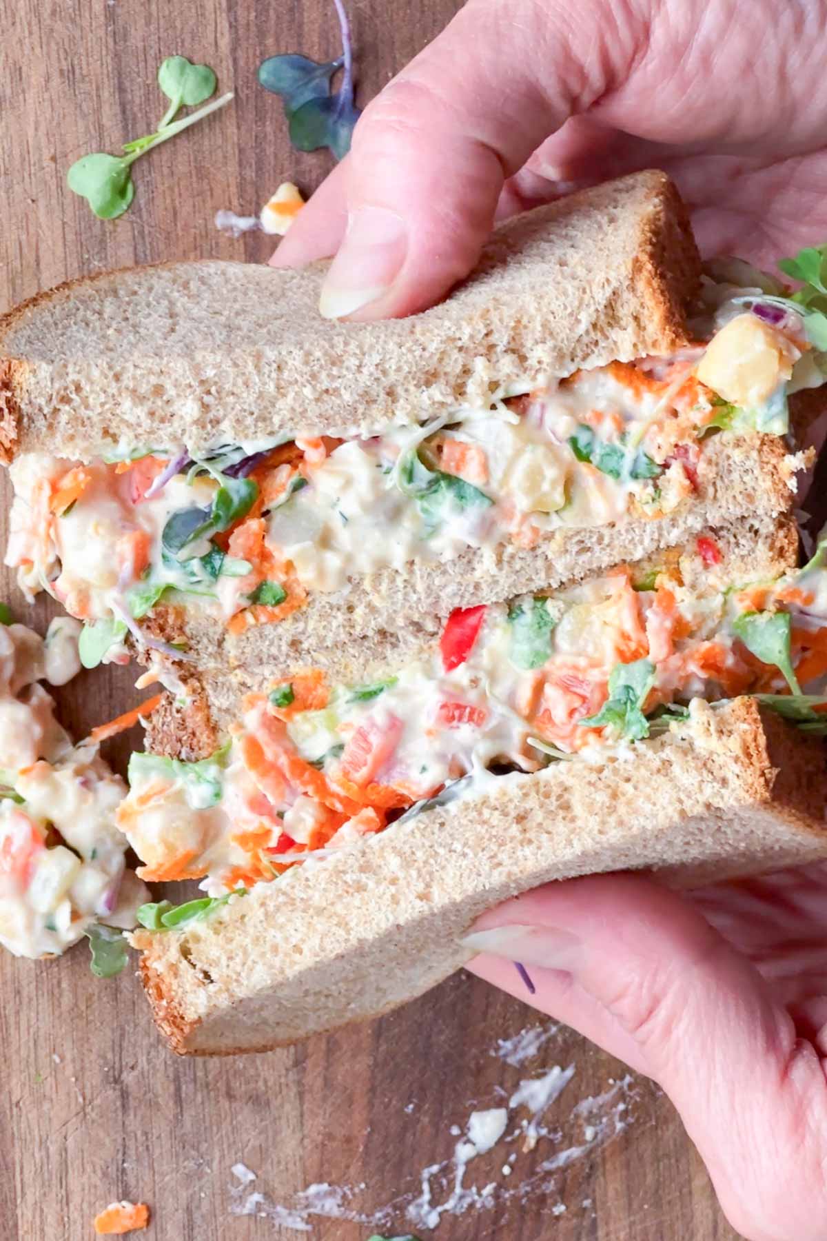 A person holds a chickpea salad sandwich made with whole wheat bread, filled with a creamy vegetable and chickpea mixture on a wooden surface.