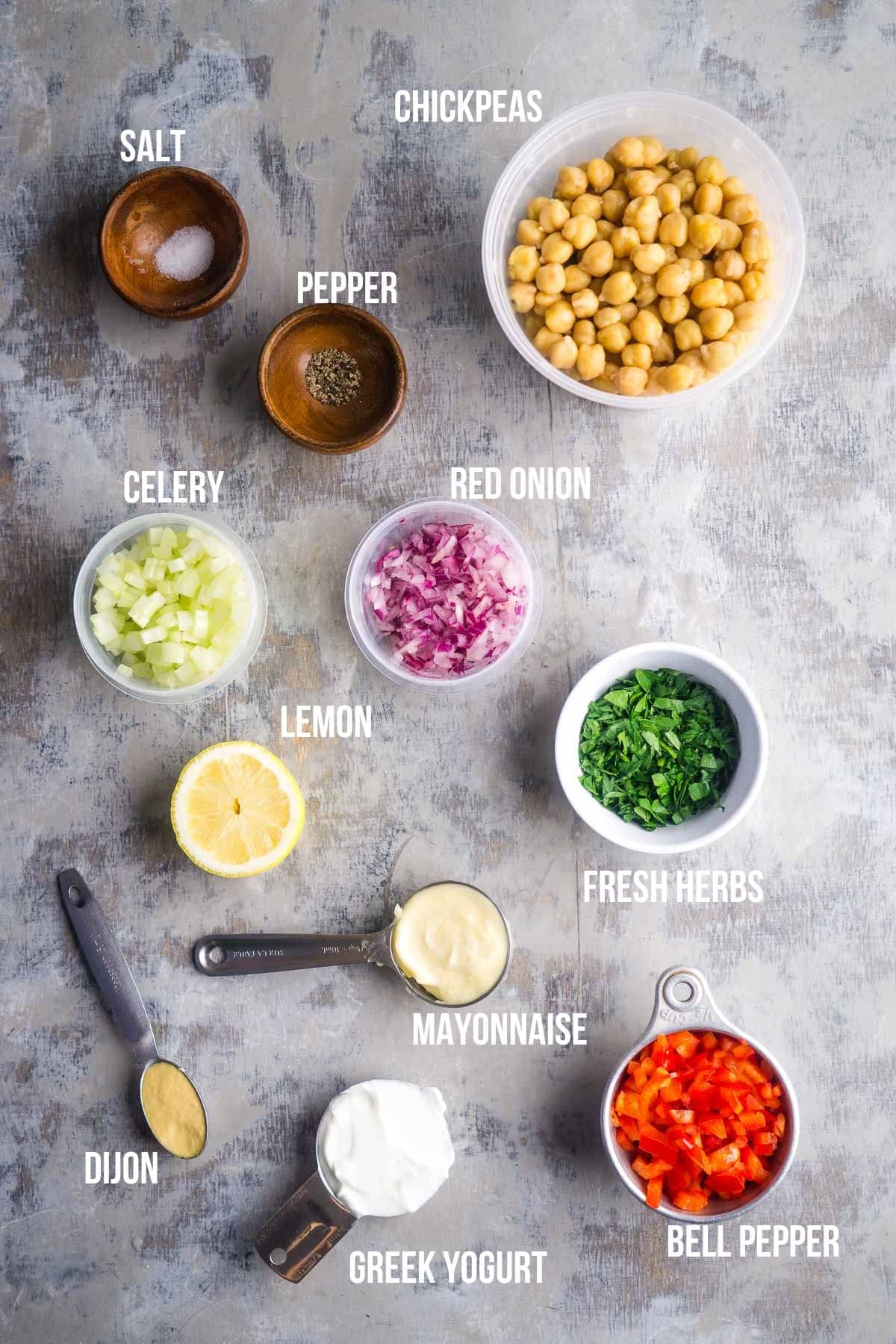 Ingredients for a chickpea salad arranged on a counter, including chickpeas, salt, pepper, celery, red onion, lemon, fresh herbs, bell pepper, Dijon, mayonnaise, and Greek yogurt.