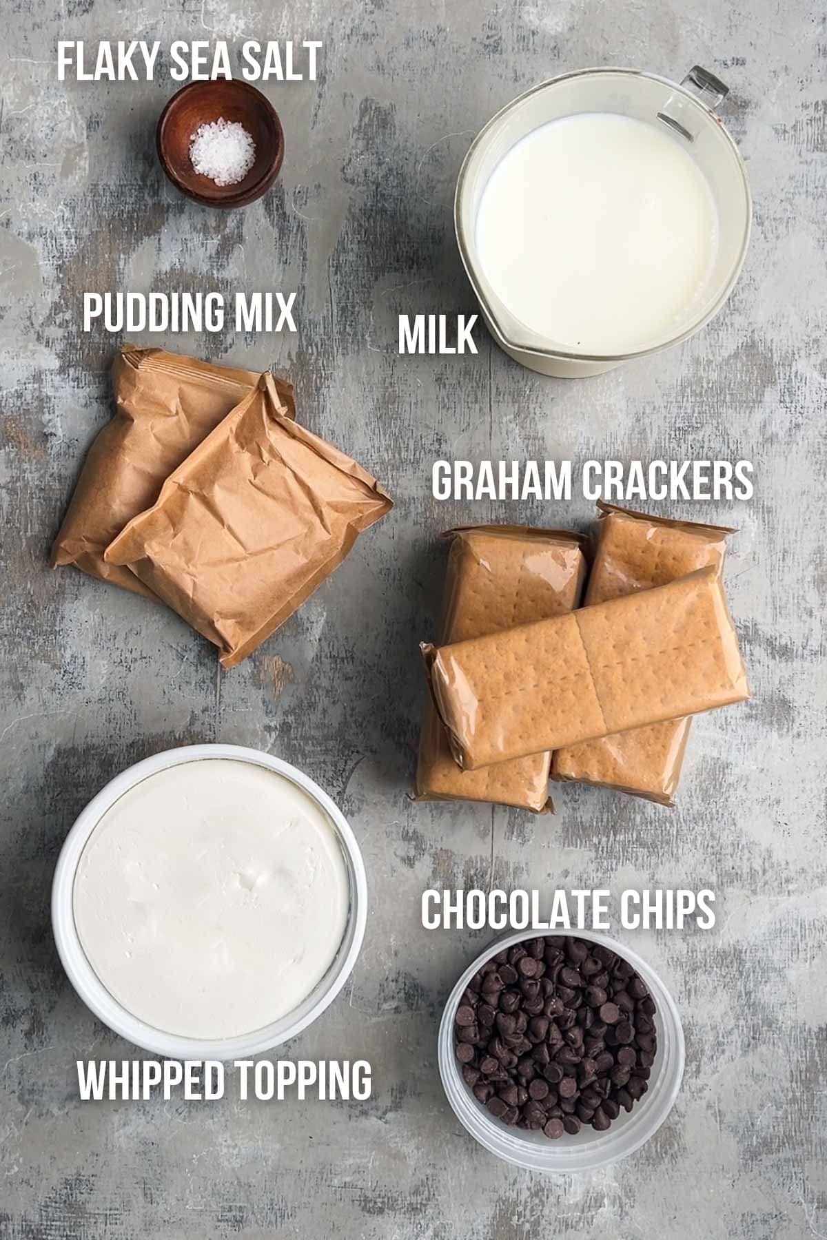Labeled ingredients on a grey surface for a choclate eclair cake: flaky sea salt, pudding mix, milk, graham crackers, whipped topping, and chocolate chips.