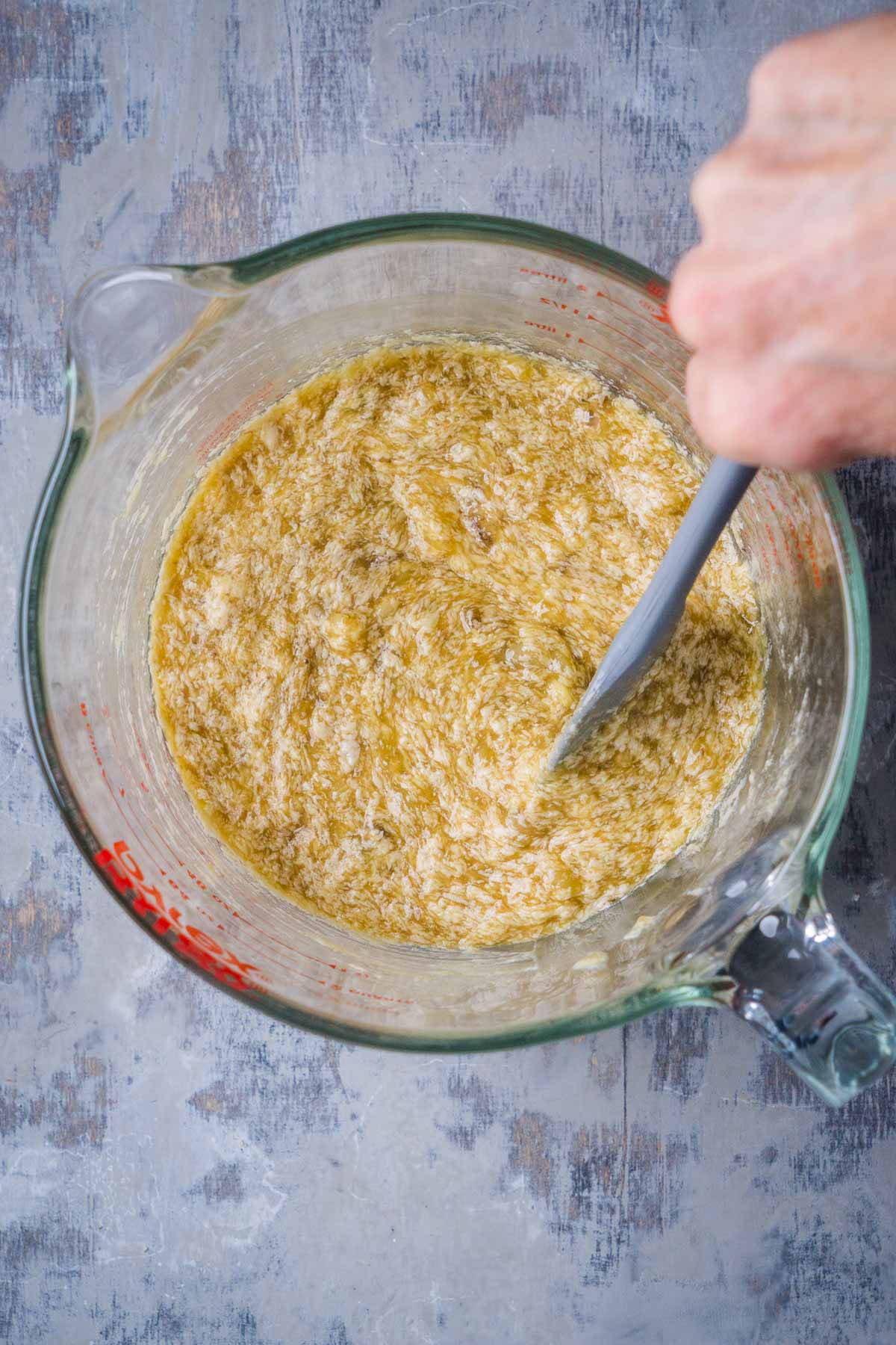 person's hand holding spatula to mix wet ingredients in glass mixing bowl