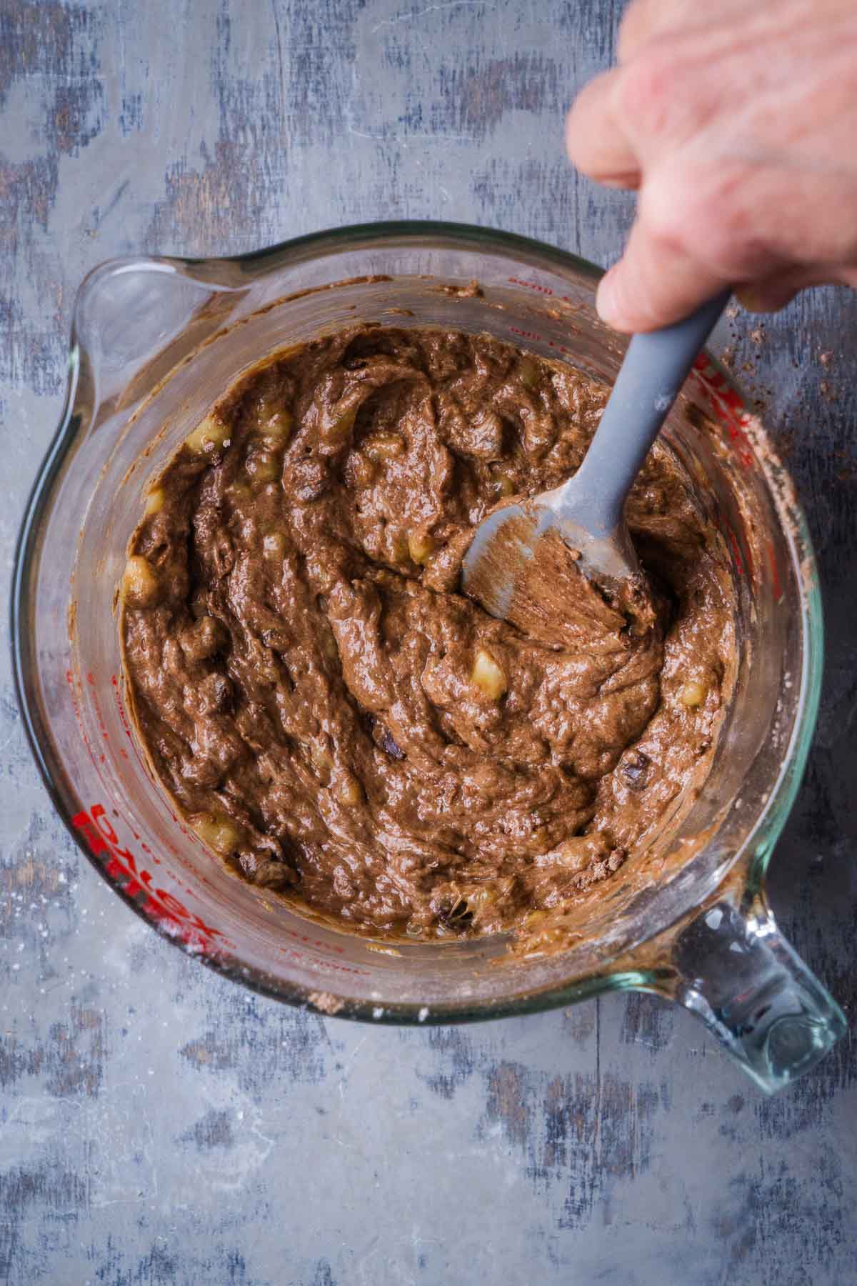 person's hand holding spatula to mix chocolate banana bread ingredients in glass mixing bowl