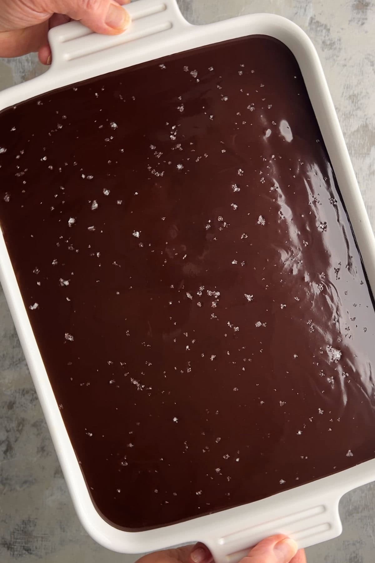 A person holding a white rectangular dish filled with smooth, glossy chocolate eclair cake topped with a sprinkling of sea salt.