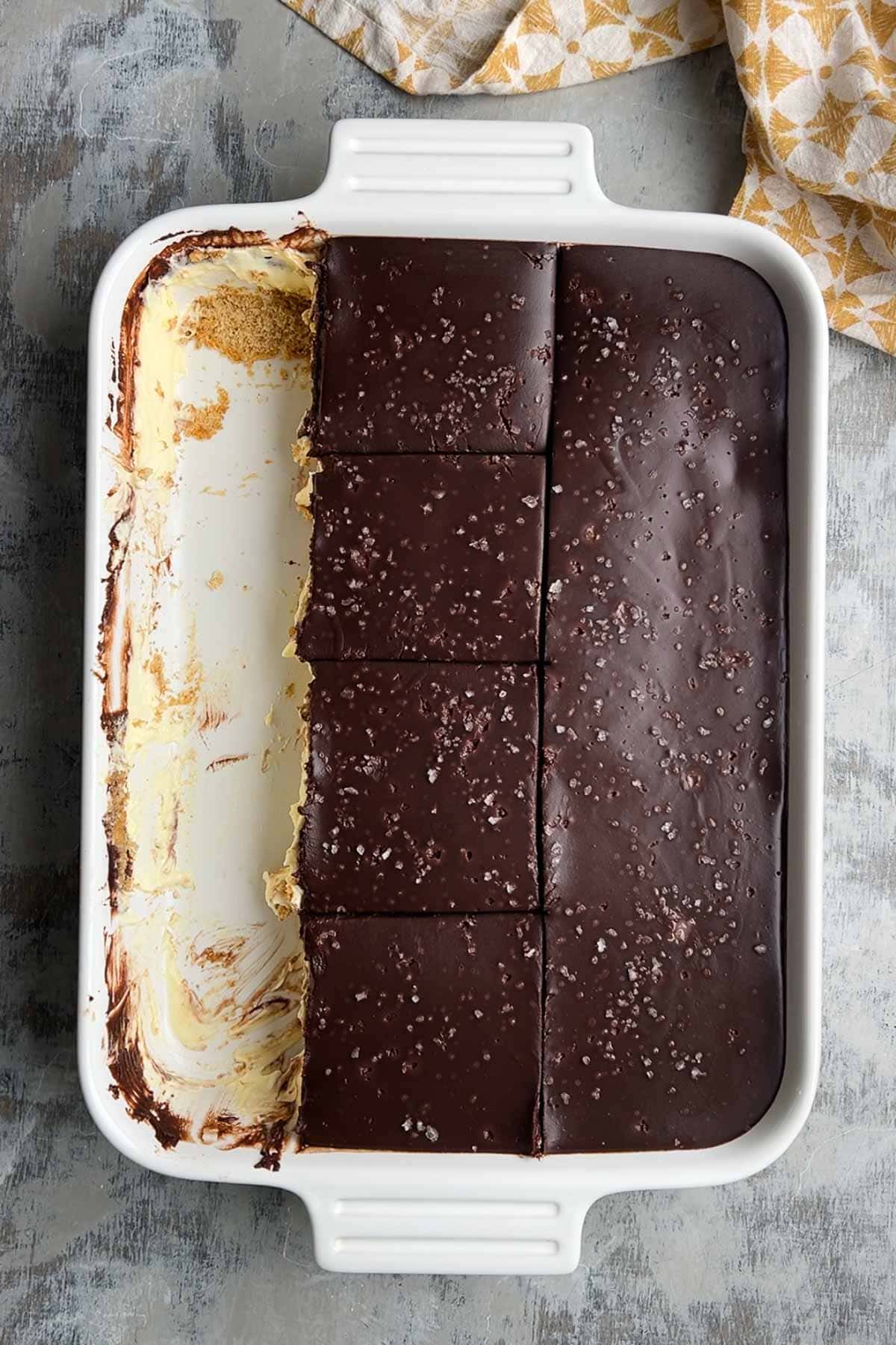 A white baking dish with chocolate eclair cake, some pieces removed to reveal layers of chocolate and filling. The surface is sprinkled with flaky salt.