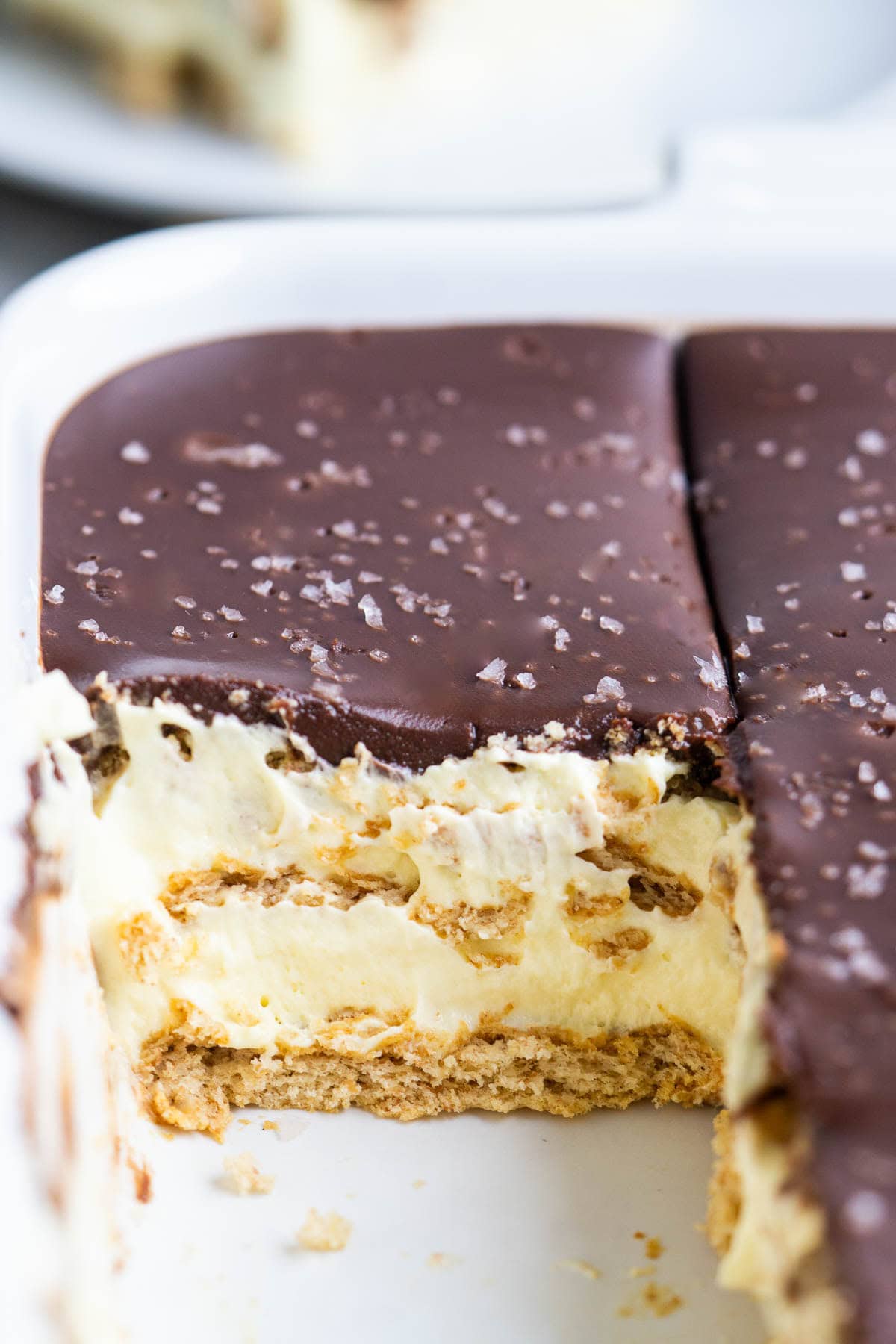 A close-up of a chocolate eclair cake with layers of graham crackers, creamy filling, and a rich chocolate topping in a white dish, with a slice removed.