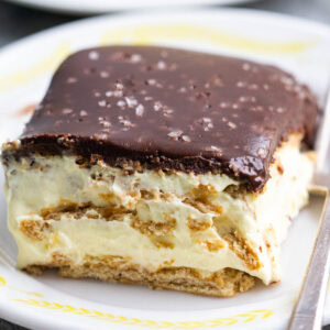 A plate of one slice of chocolate eclair cake with a graham cracker base, creamy filling, and a thick chocolate topping, garnished with flaky salt. A fork rests on the plate.
