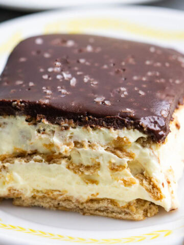 A plate of one slice of chocolate eclair cake with a graham cracker base, creamy filling, and a thick chocolate topping, garnished with flaky salt. A fork rests on the plate.