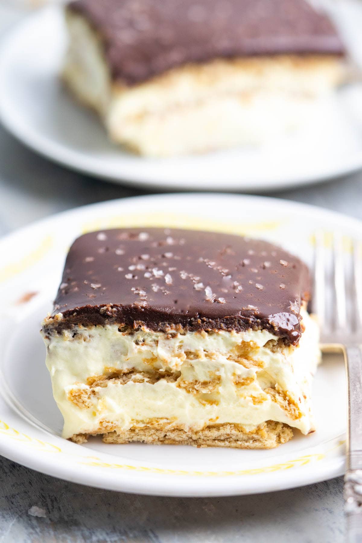 Chocolate eclair cake is a crowd-favorite no-bake dessert made with layers of graham crackers, creamy vanilla filling, and chocolate topping. This chilled dessert is easy to make ahead and perfect for feeding a crowd because the refrigerator does most of the work. Two plates, each with one slice of chocolate eclair cake with a graham cracker base, creamy filling, and a thick chocolate topping, garnished with flaky salt. A fork rests on the plate.