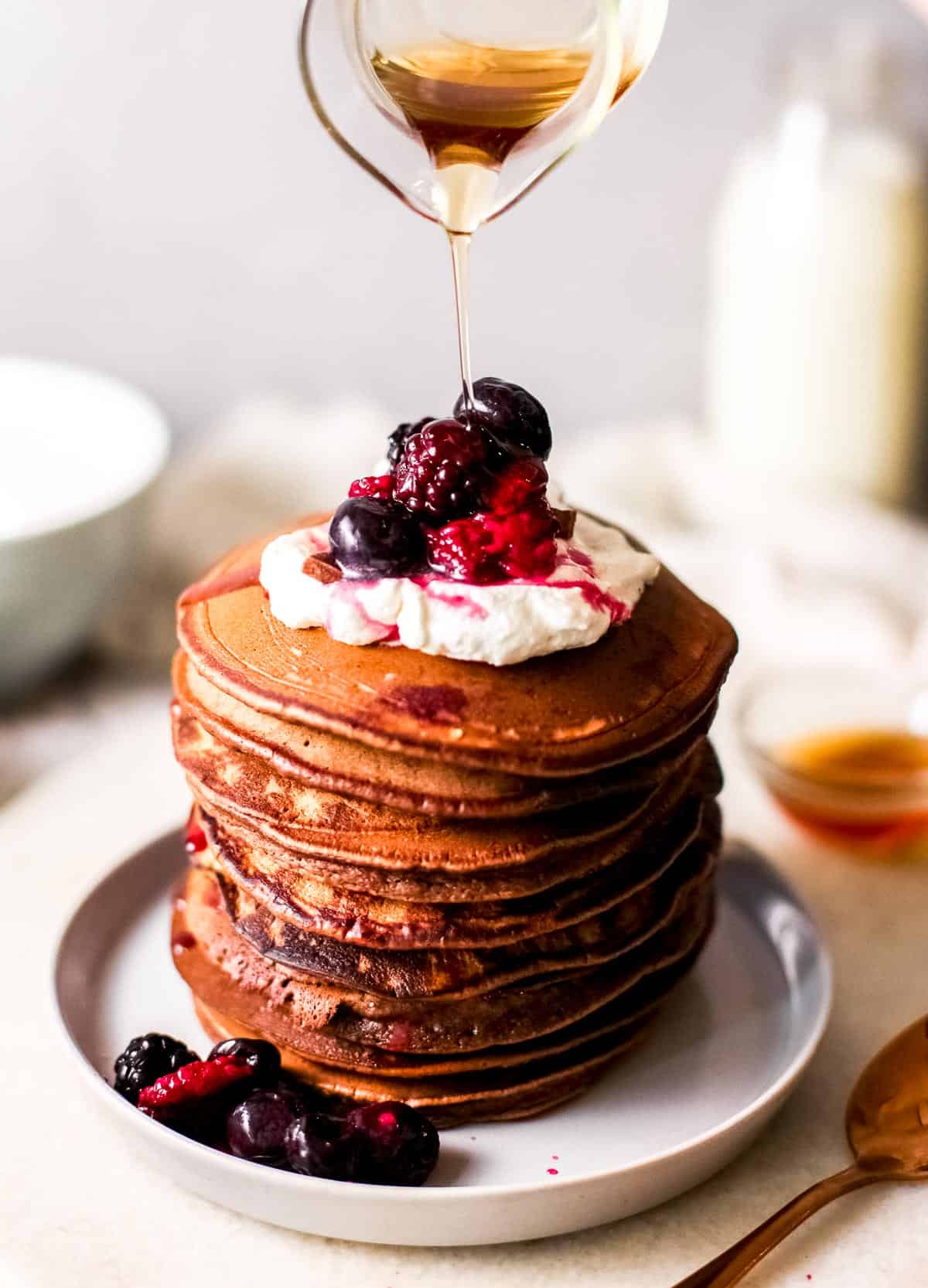 syrup pouring over a stack of chocolate pancakes topped with whipped cream and fresh berries