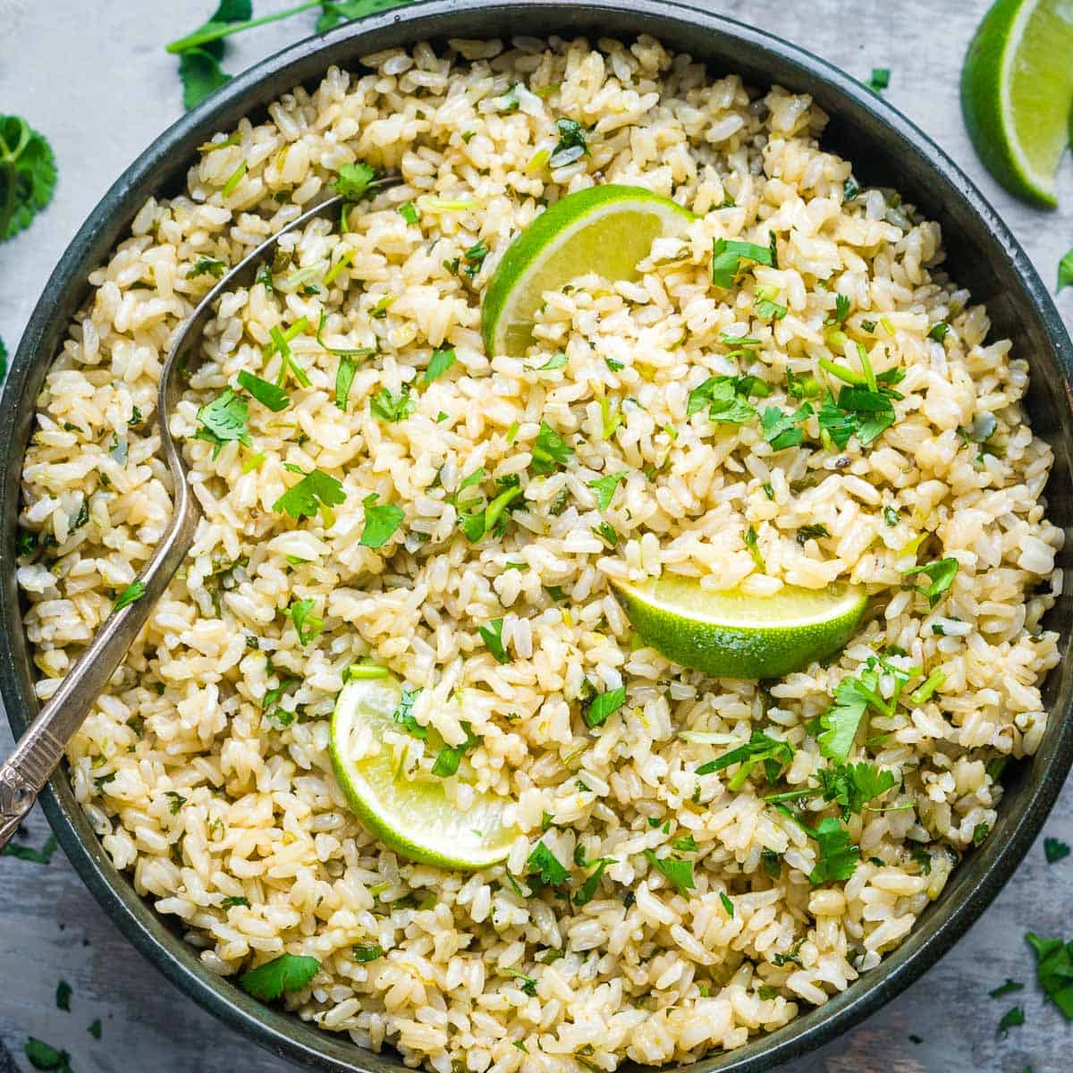 cilantro lime brown rice in serving bowl with serving spoon garnished with lime wedges and chopped cilantro