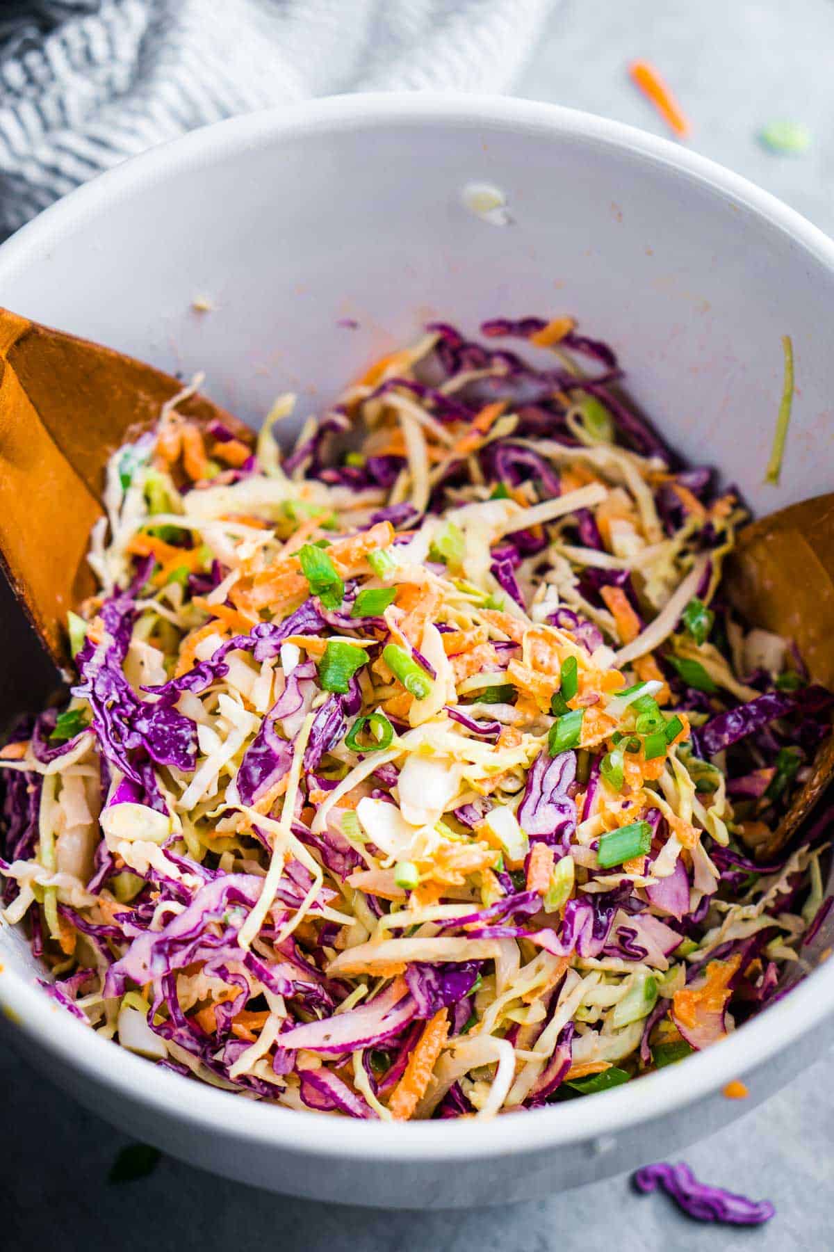 coleslaw ingredients in white bowl being tossed by wooden salad tongs