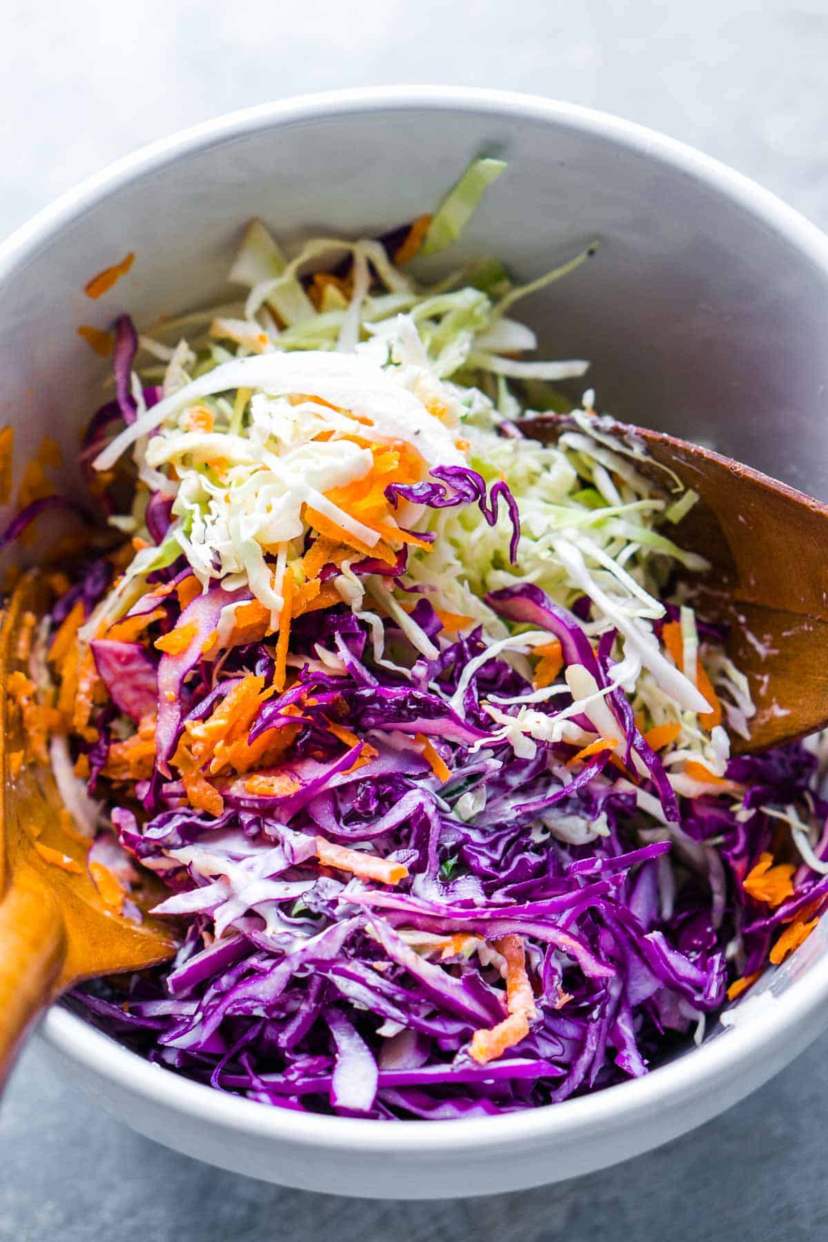 coleslaw salad ingredients in white bowl are tossed with wood salad utensils