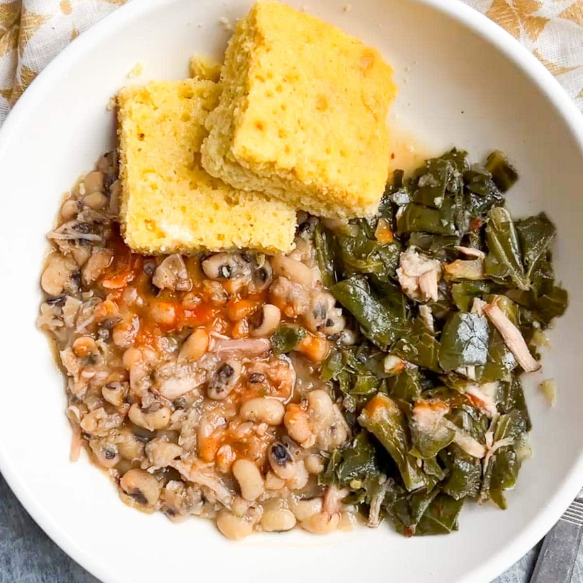 A white bowl of black-eyed peas with hot sauce, savory collard greens with pieces of turkey, and two squares of cornbread on top.