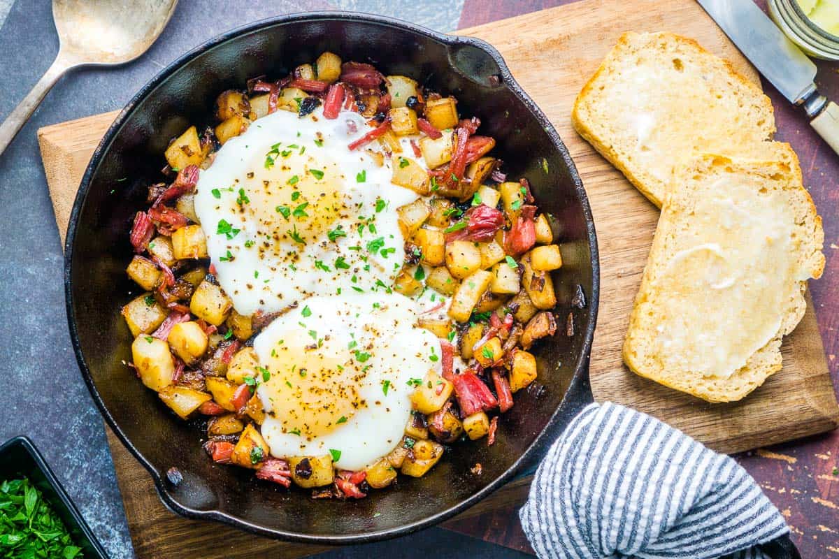 eggs over corned beef hash in iron skillet on board with toasted beer bread slices