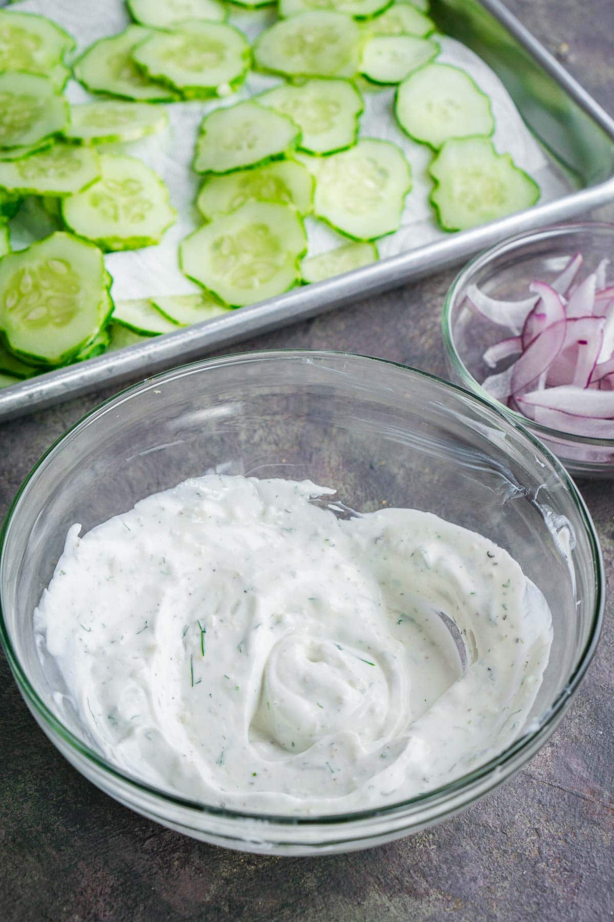 A glass bowl of creamy white dressing sits beside a bowl of sliced red onions and a tray of sliced cucumbers on a kitchen counter.