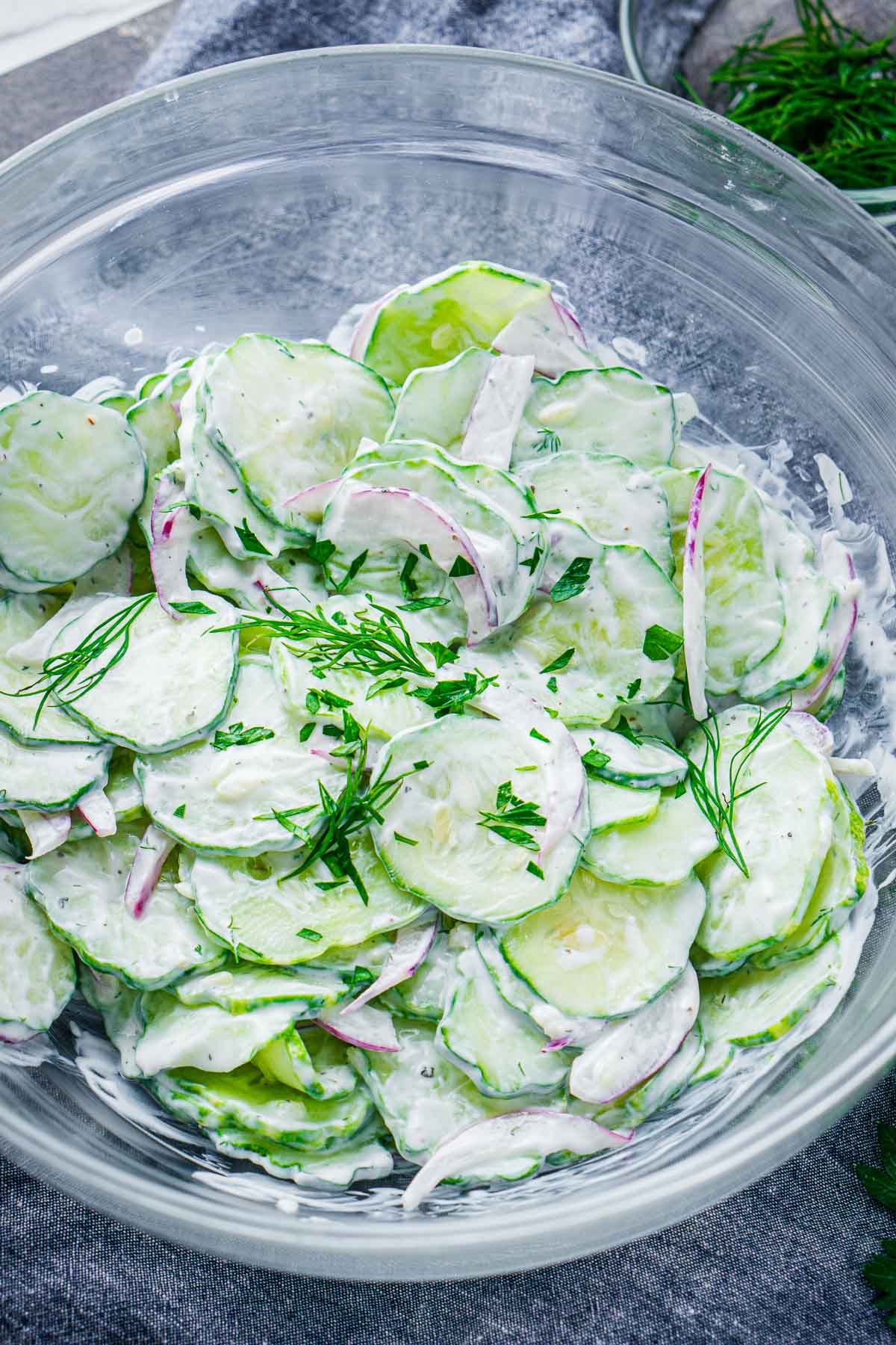 A glass bowl filled with creamy cucumber salad, featuring sliced cucumbers, red onions, and fresh dill on top.