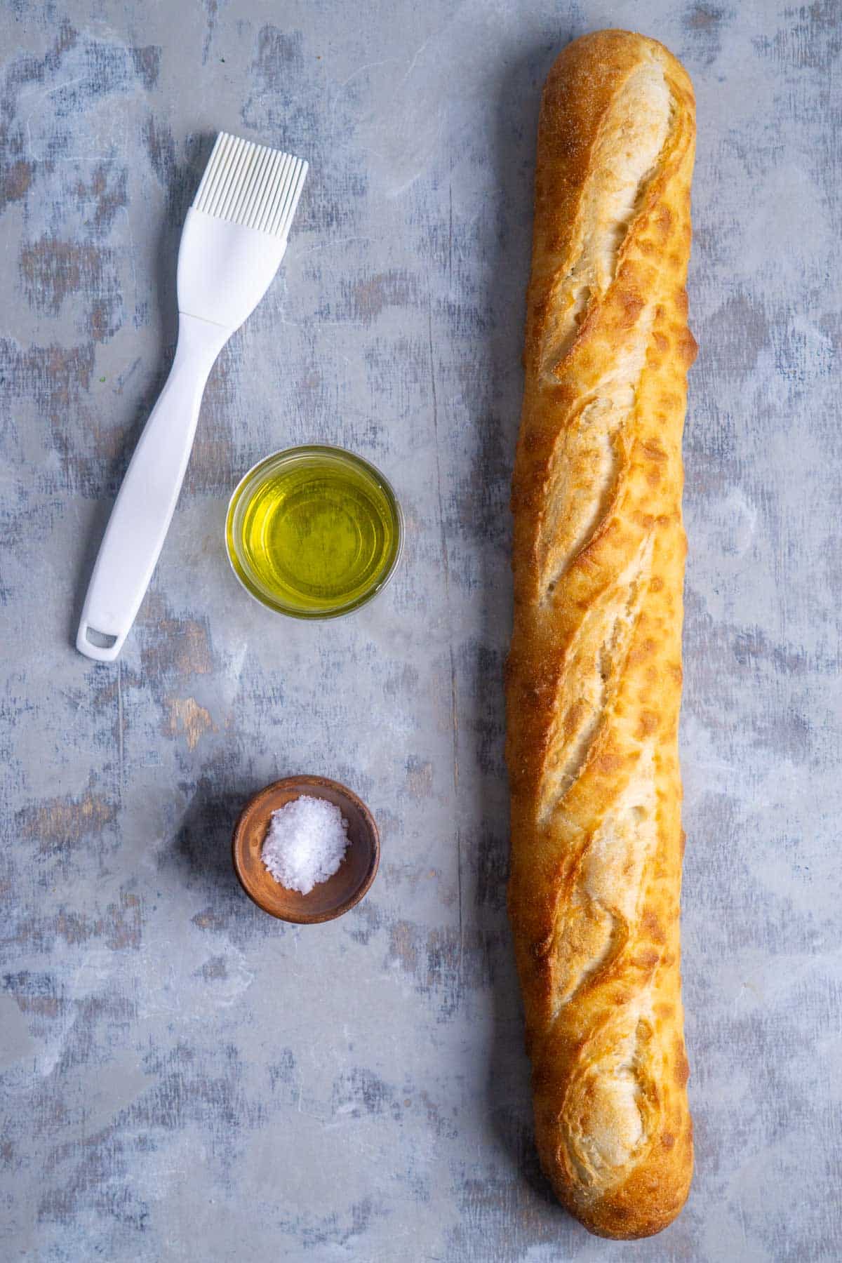 whole baguette next to glass jar of olive oil, wood bowl of salt, and white silicone pastry brush