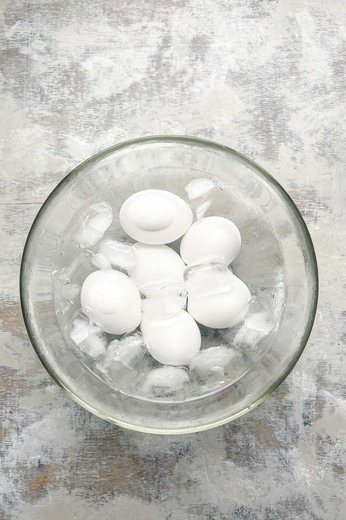 A glass bowl filled with ice cubes and six white eggs, viewed from above, on a textured gray surface.