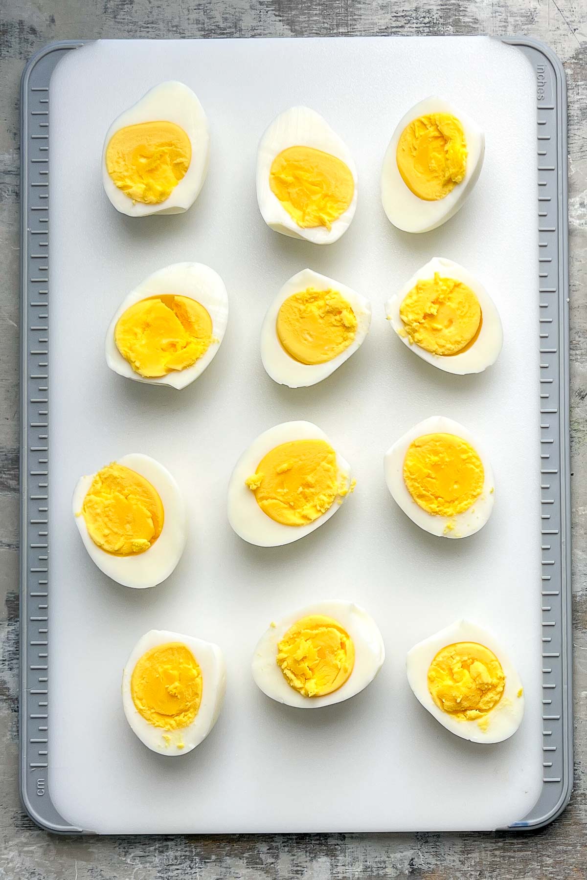 Twelve hard-boiled eggs, sliced in half are arranged in rows on a white cutting board.