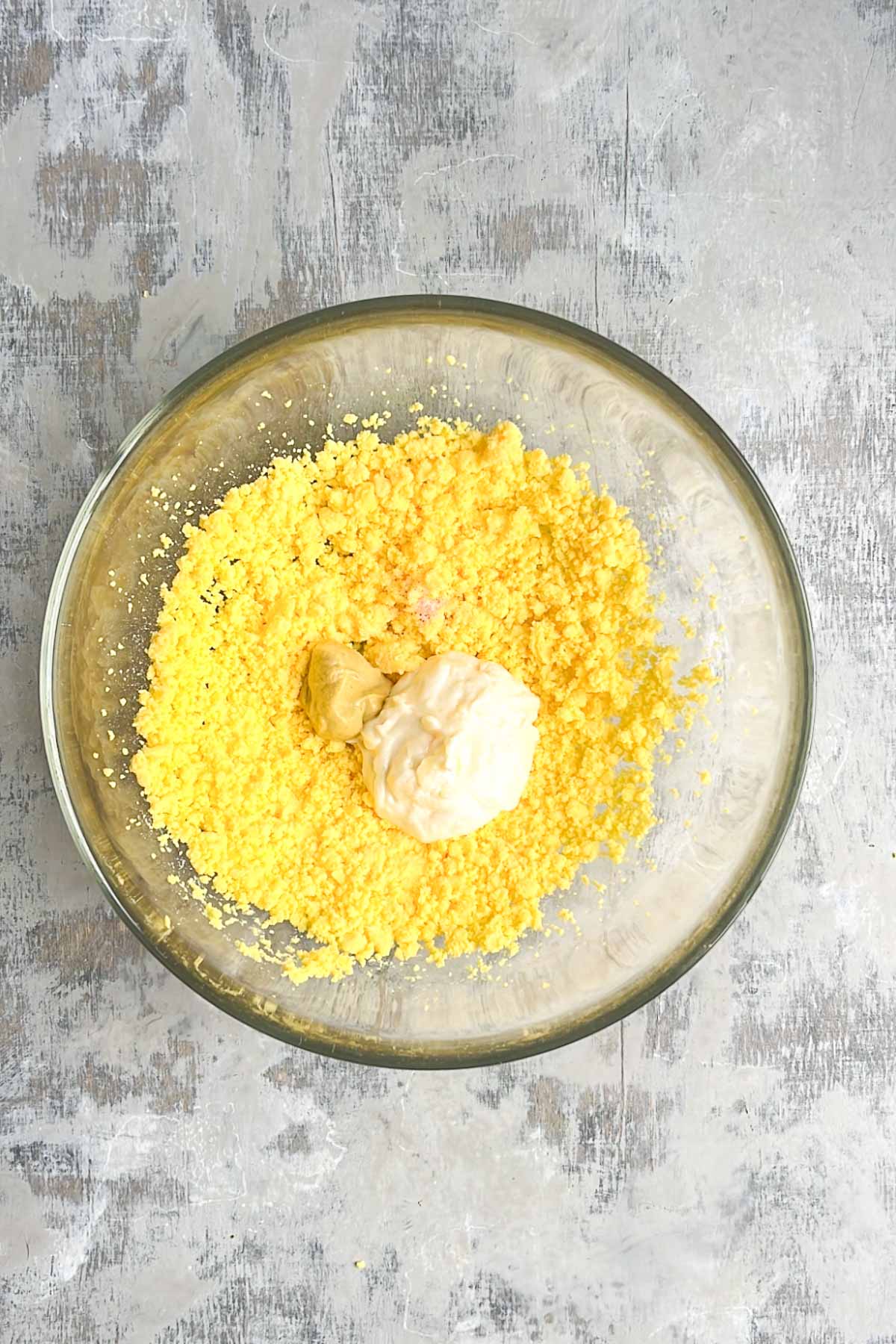 A glass bowl containing crumbled hard-boiled egg yolks, a dollop of mayonnaise, and a dollop of mustard.