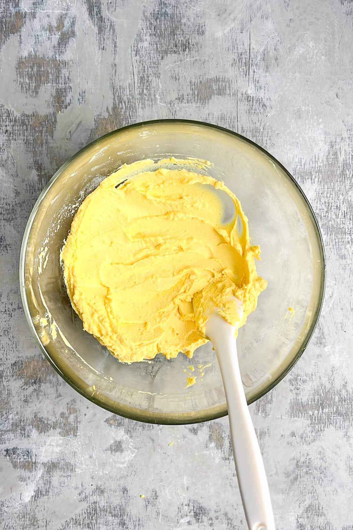 A glass bowl filled with yellow filling for deviled eggs, with a white spatula resting inside.
