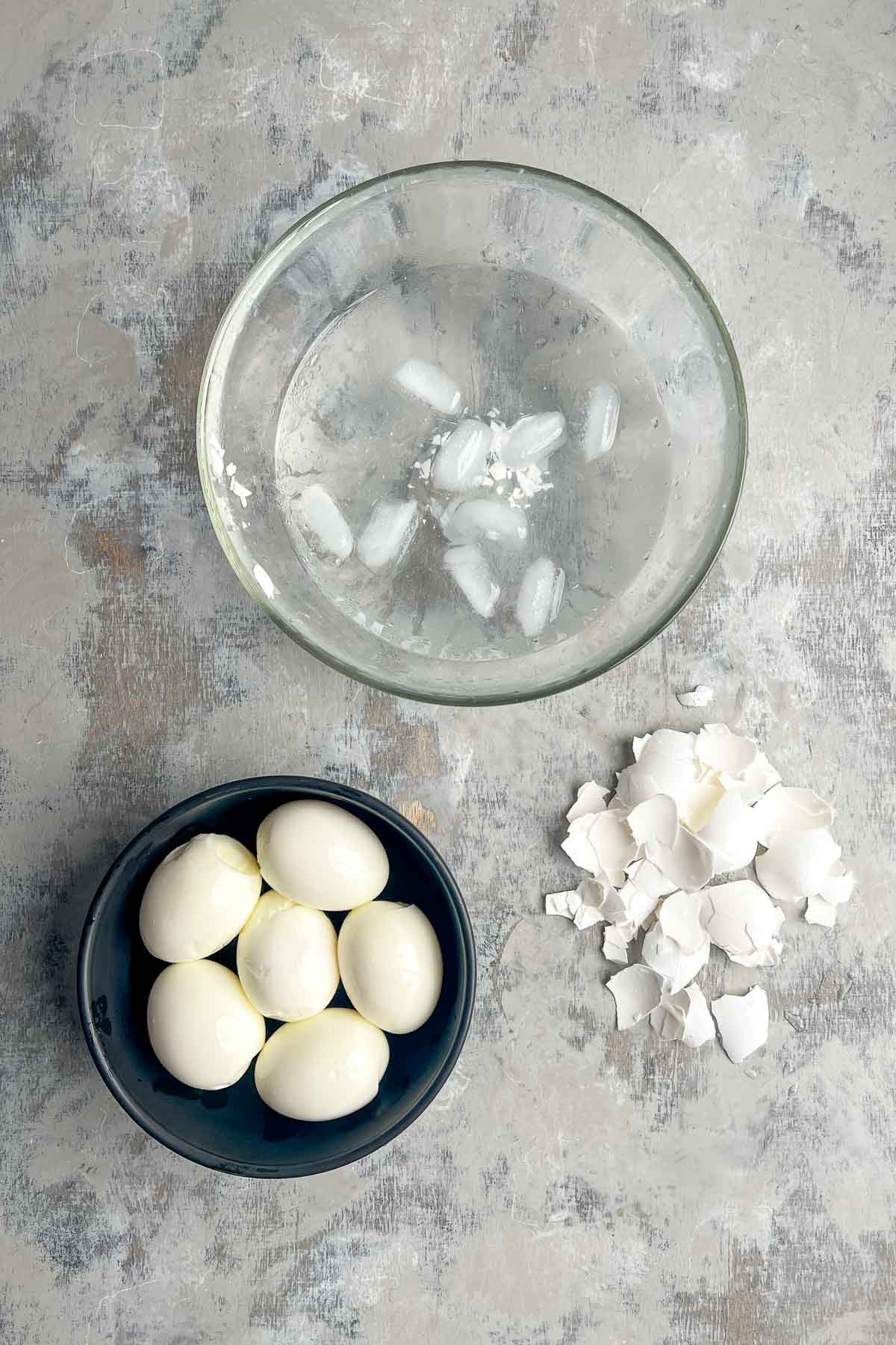 A bowl of ice water, a bowl of peeled hard-boiled eggs, and a pile of eggshells are arranged on a gray surface.