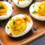 Several deviled eggs on a wooden tray, topped with paprika and chopped chives, make a classic appetizer everyone will love.