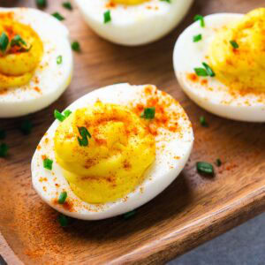 Several deviled eggs on a wooden tray, topped with paprika and chopped chives, make a classic appetizer everyone will love.