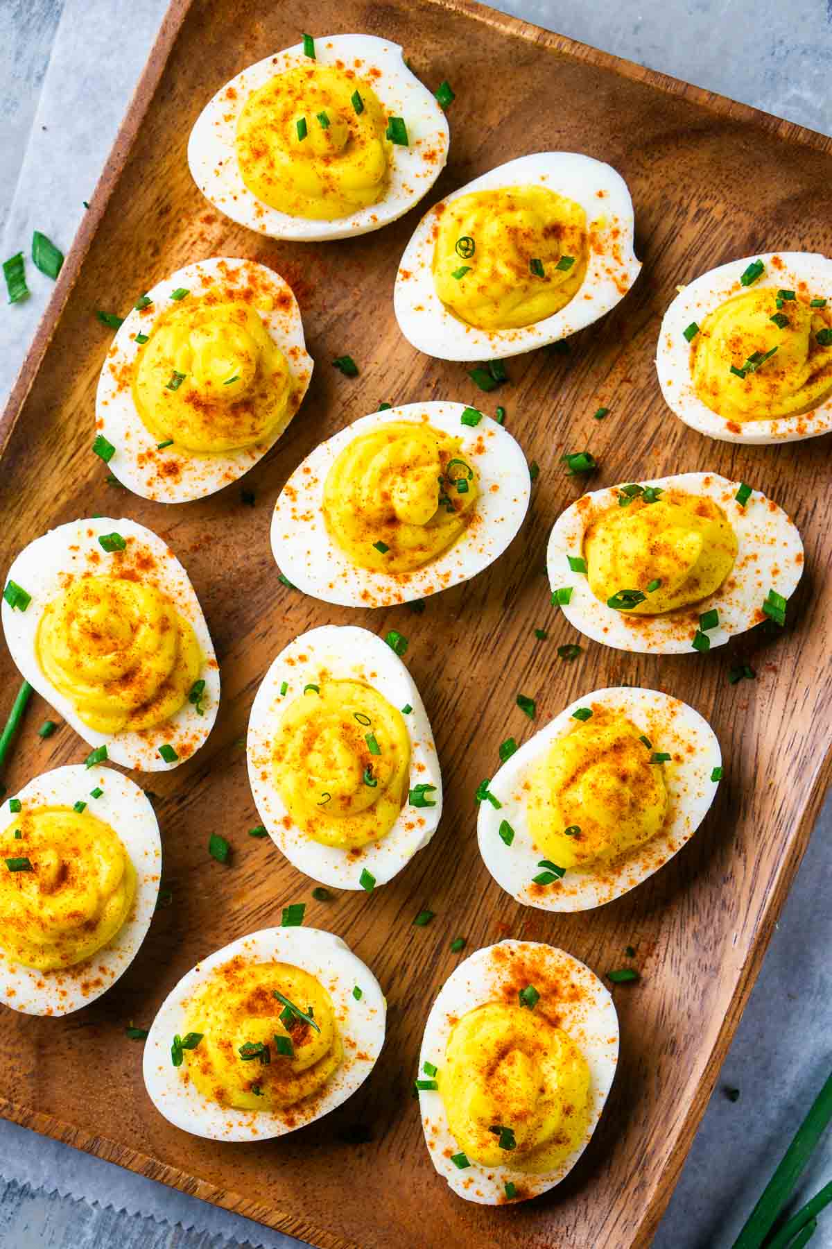 A wooden tray holds twelve deviled eggs, topped with paprika and chopped chives, neatly arranged in rows on a gray surface.