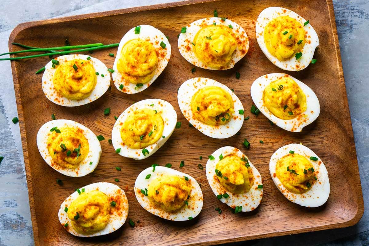 A wooden tray holds twelve deviled eggs, topped with paprika and chopped chives, neatly arranged in rows on a gray surface.