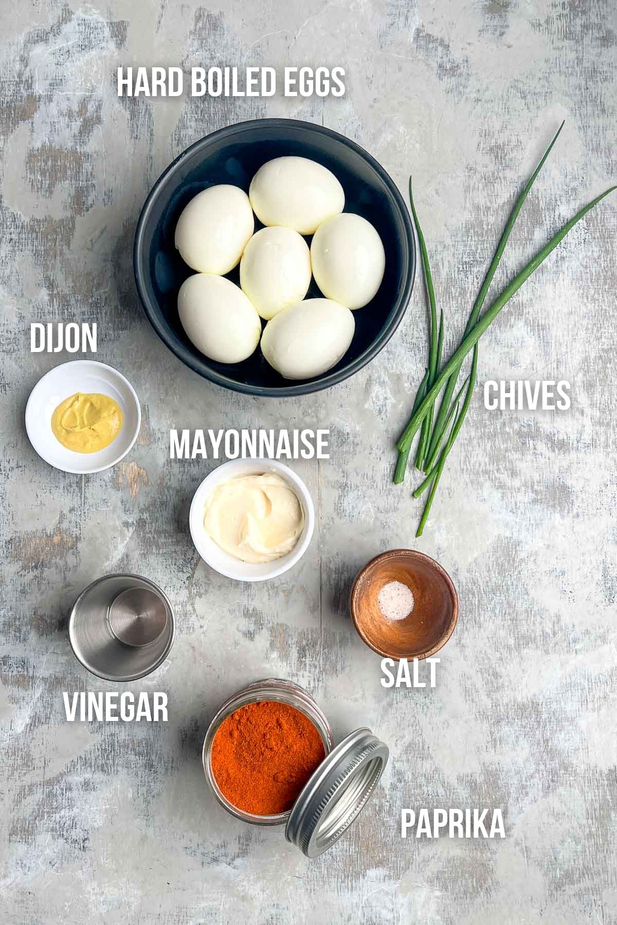 Bowl of hard boiled eggs for deviled eggs, chives, small dishes of Dijon mustard, mayonnaise, vinegar, salt, and a jar of paprika arranged on a gray surface, each labeled.