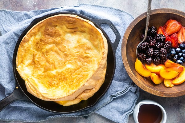 dutch baby pancake in iron skillet on blue linen next to wooden bowl of fresh fruit and white maple syrup container