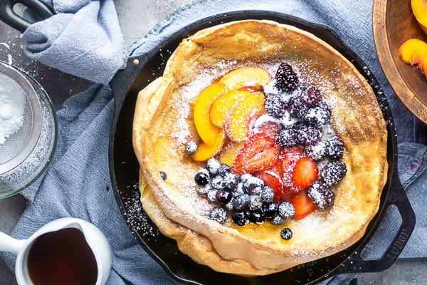 Fruit topped Dutch baby pancake in iron skillet on blue linen next to bowls of fruit, maple syrup, and powdered sugar toppings